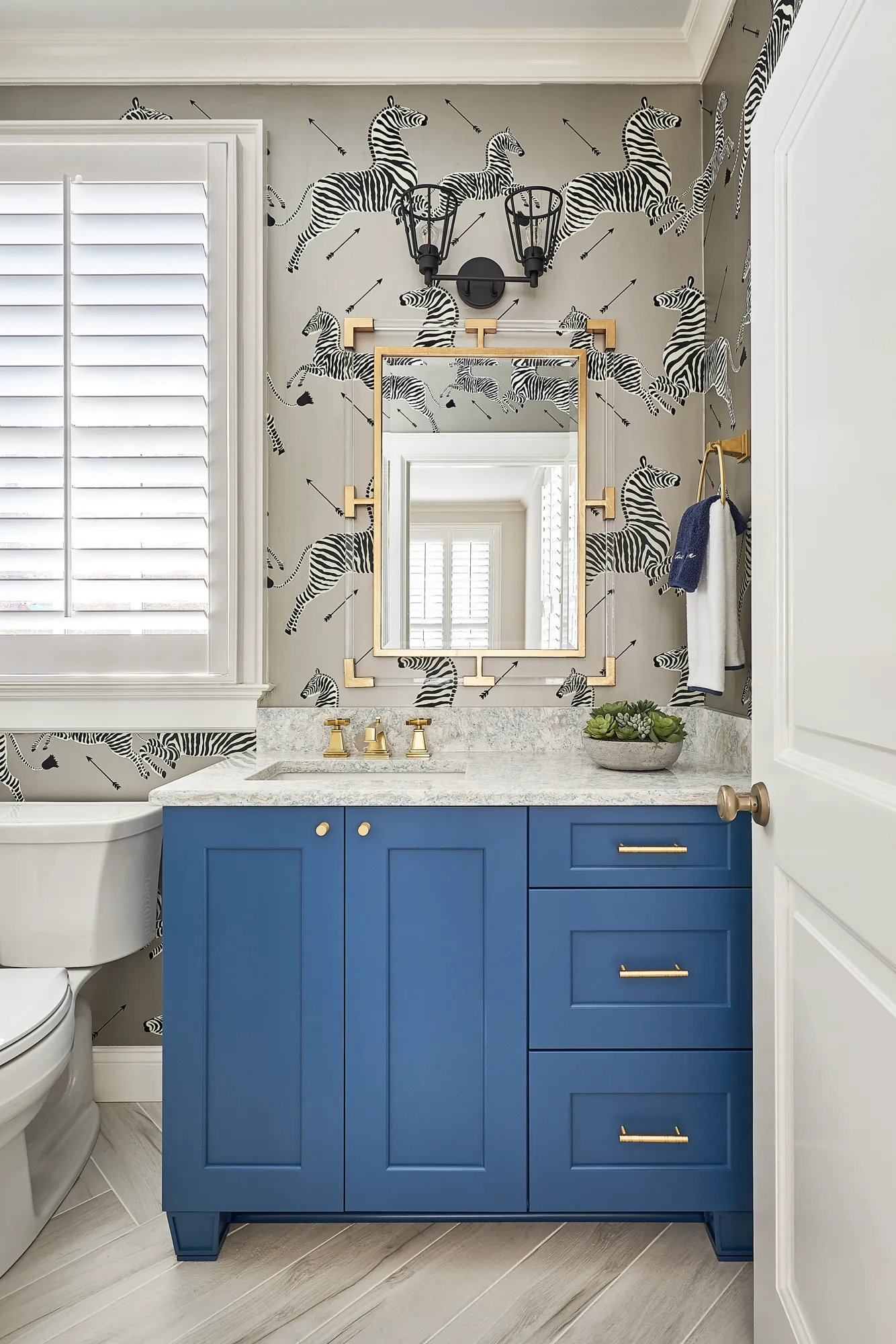 A powder room with bold zebra wallpaper and gray flooring. A deep blue vanity with a gray marble counter features gold hardware and a geometric gold mirror.