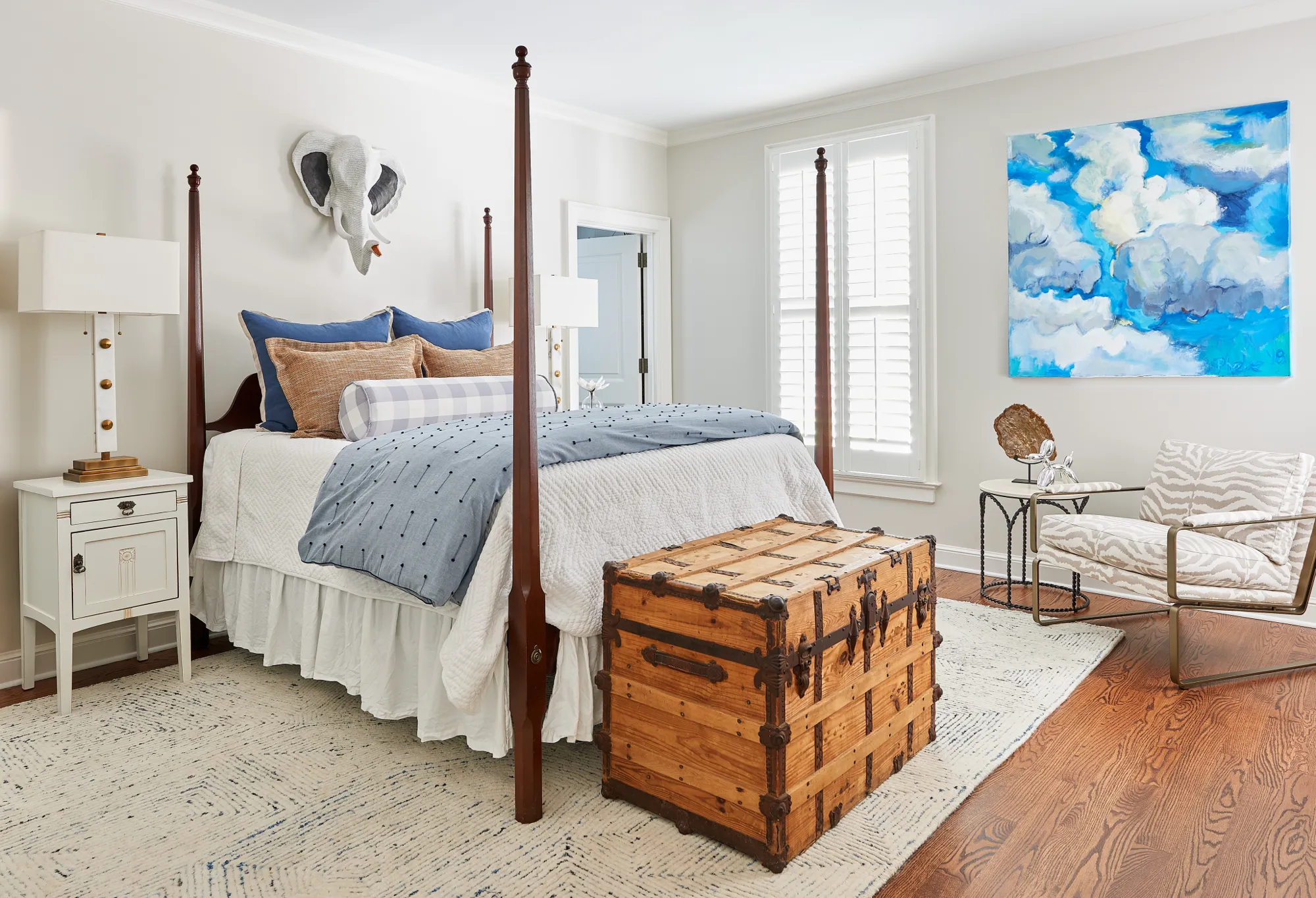 A bright transitional bedroom featuring a four-poster bed, a vintage wooden trunk, and blue and white bedding. The decor includes an elephant head wall mount and abstract cloud art.