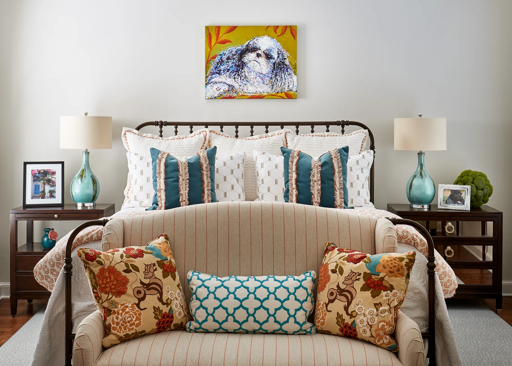 A vibrantly decorated bedroom featuring a metal bed layered with patterned pillows, flanked by turquoise lamps. A striped loveseat with floral cushions sits at the foot of the bed.