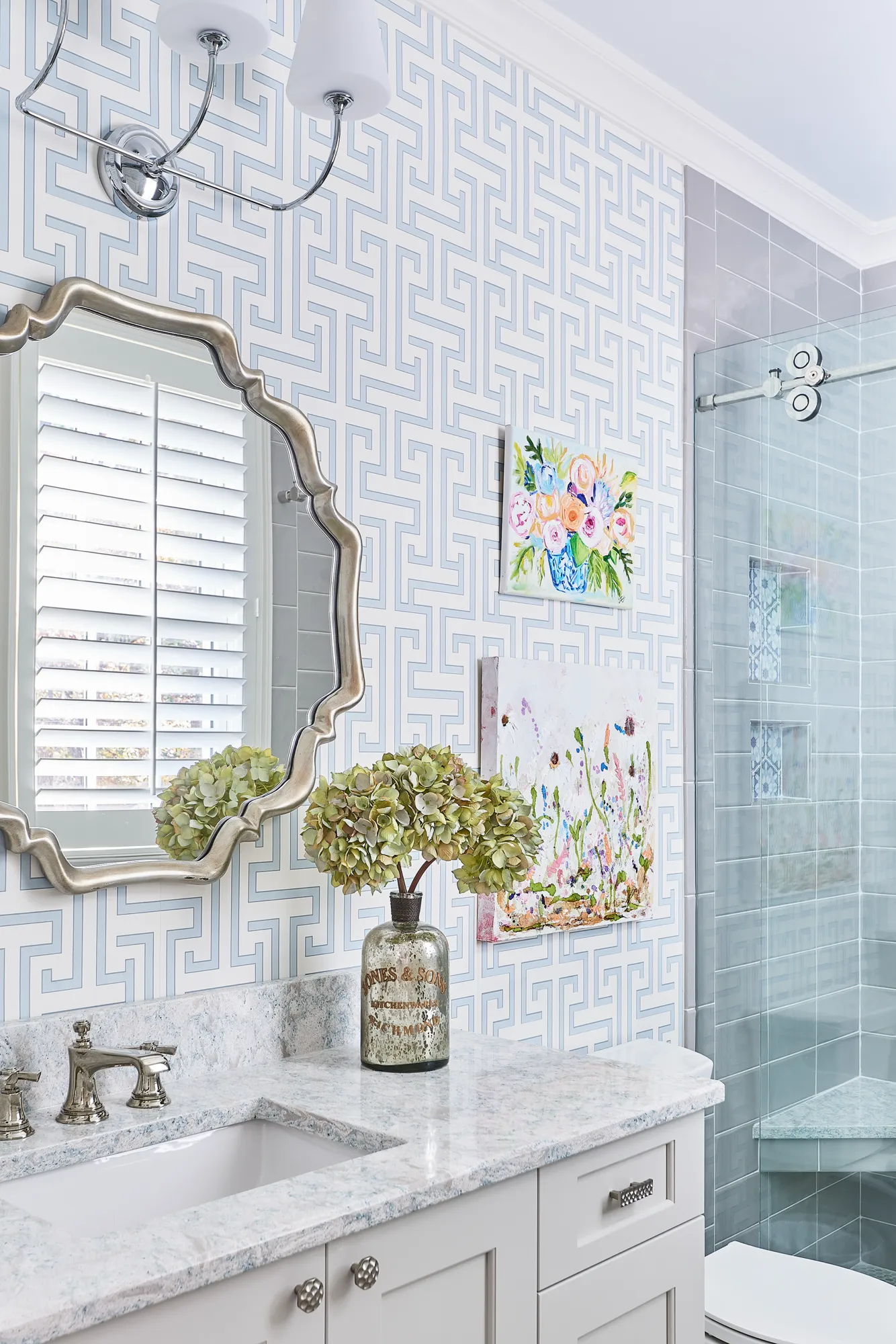 A stylish bathroom vanity with a marble countertop, silver fixtures, and an ornate mirror. The walls feature light blue Greek key wallpaper contrasting with the tiled shower.