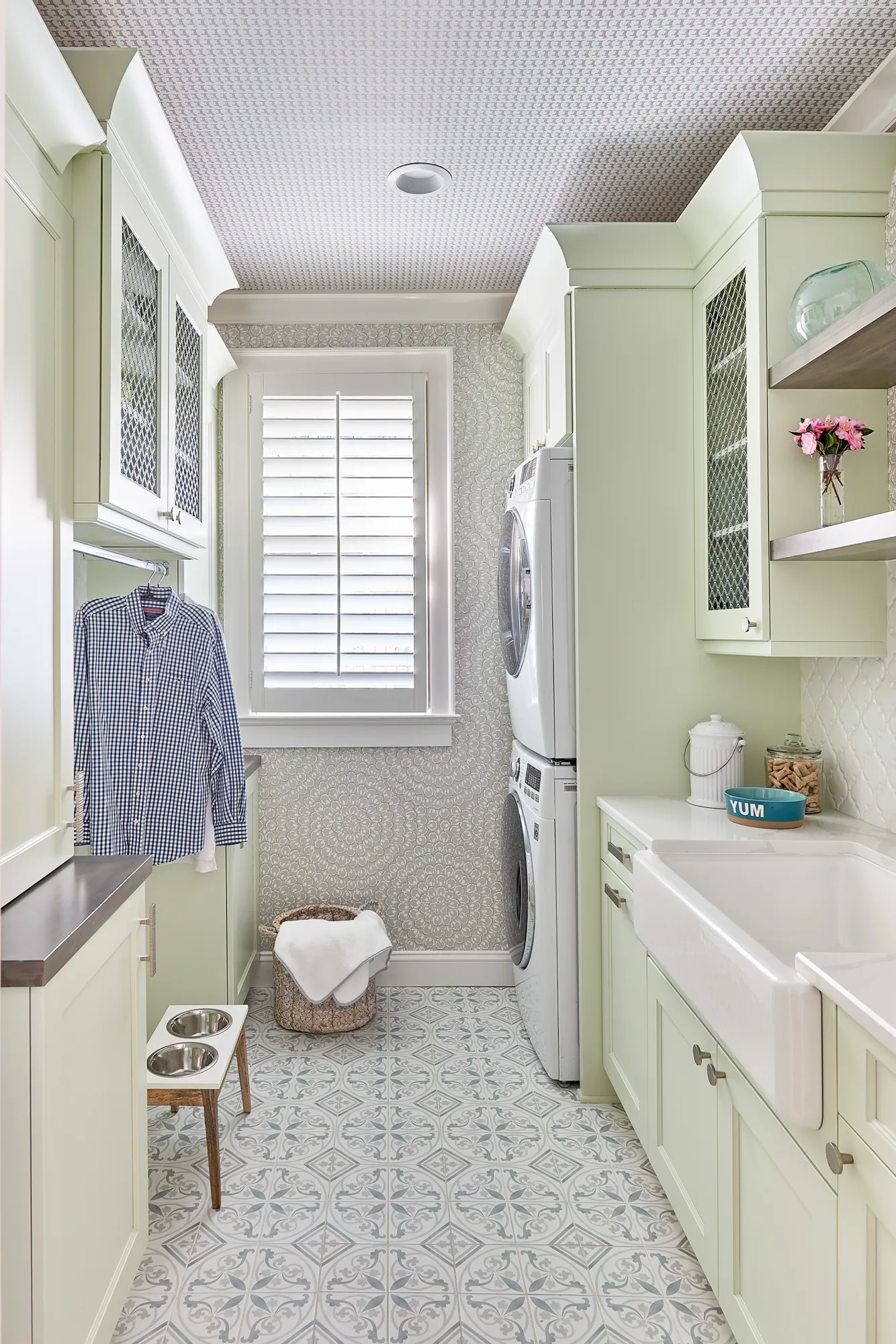 A bright laundry room with light green cabinetry, patterned tile floor, a stacked washer/dryer, and a large farmhouse sink. A hanging shirt and dog bowls add function.