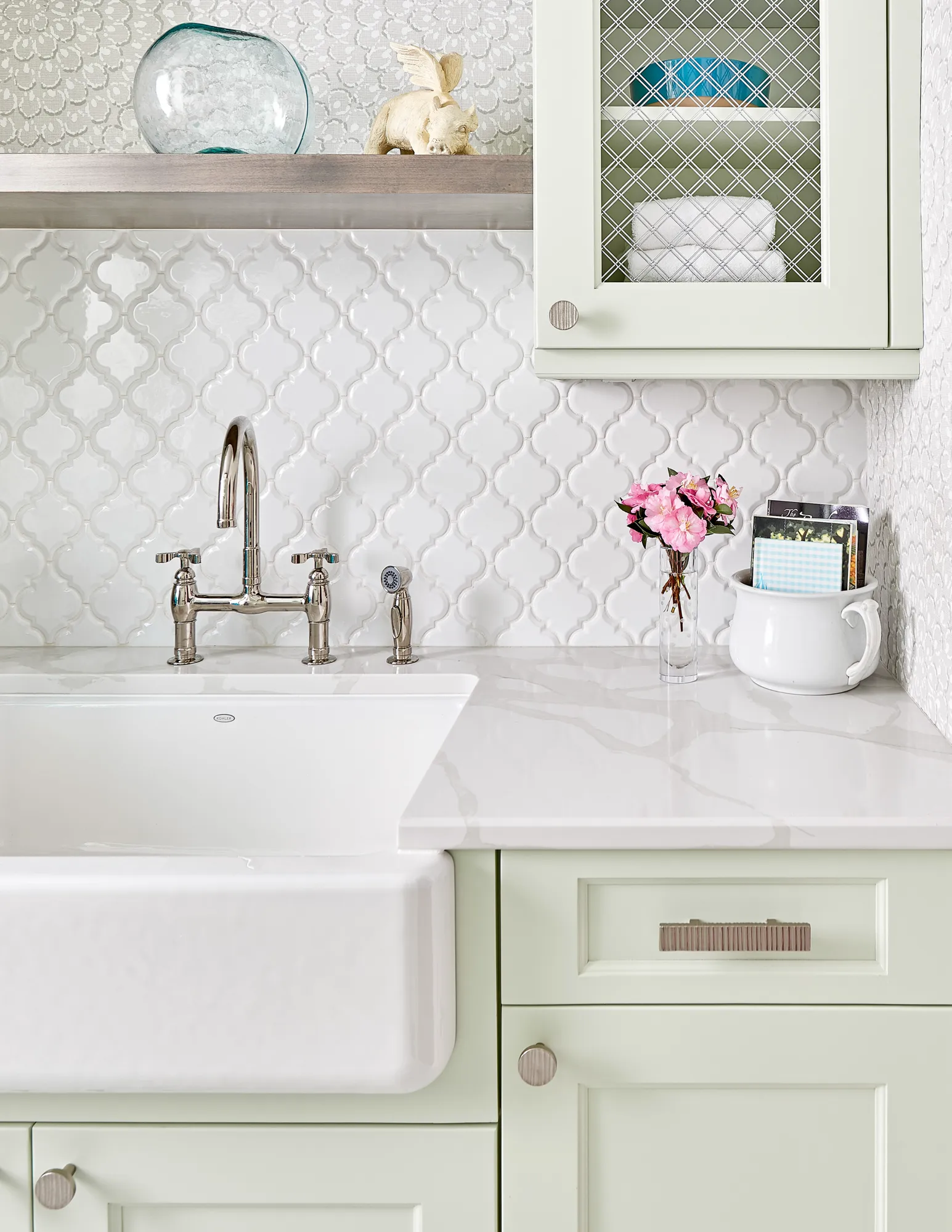 A bright utility area featuring a white apron-front sink, chrome bridge faucet, and light green cabinets with a white arabesque tile backsplash. Decor includes pink flowers.
