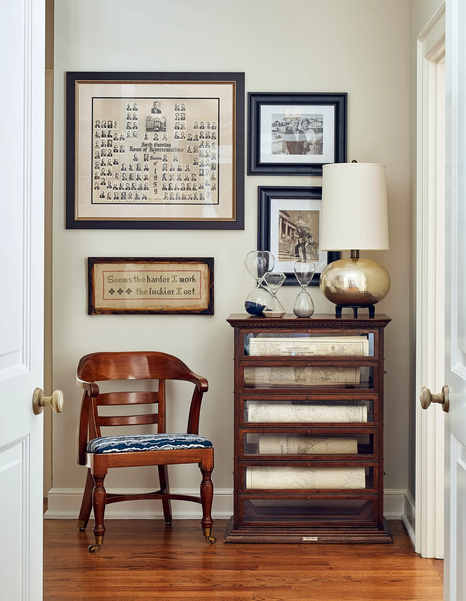 A gallery wall of framed vintage prints and a cross-stitch sampler overlooks a wooden armchair and antique flat file cabinet displaying rolled maps, hourglasses, and a gold lamp.