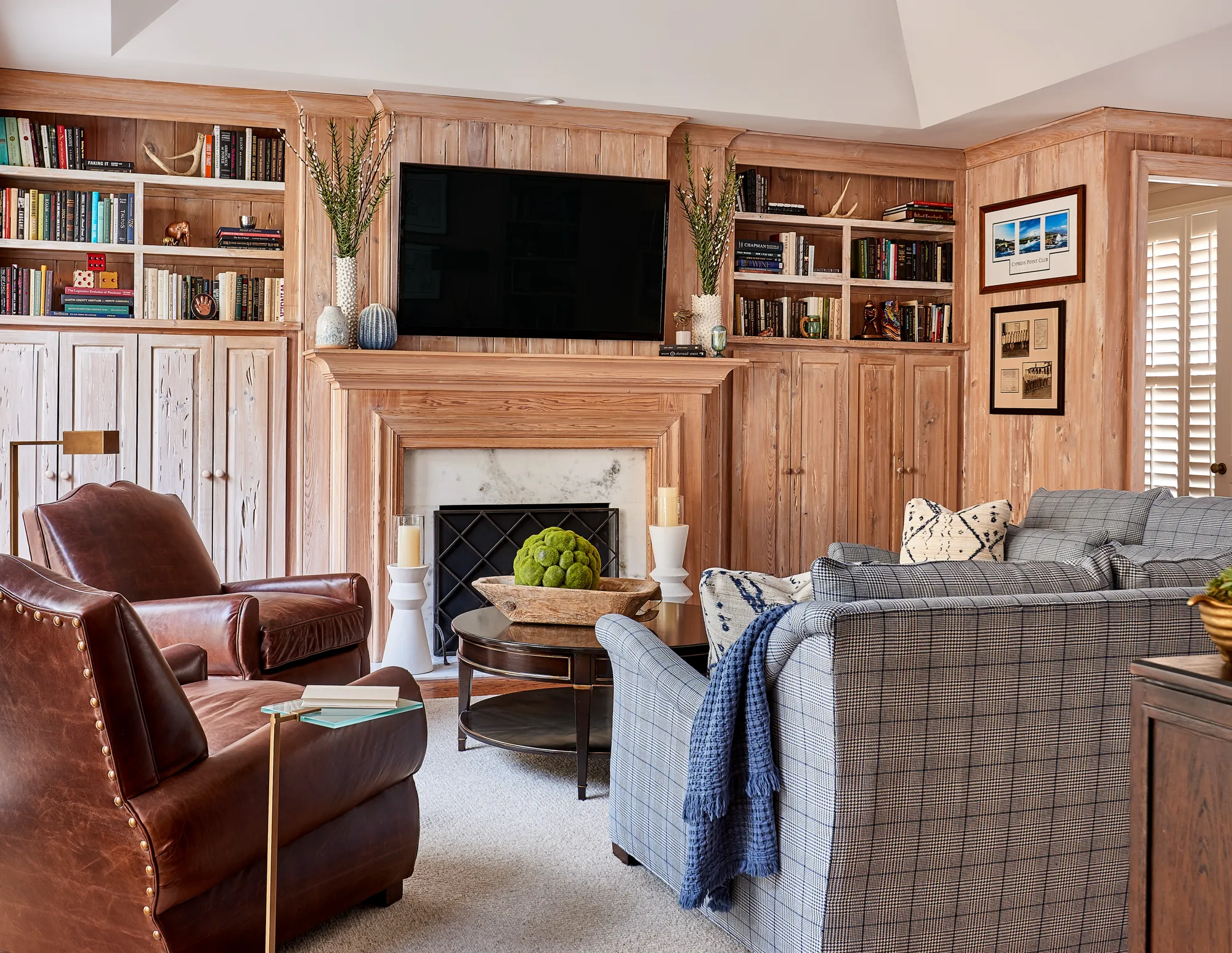 A cozy living room featuring walls clad in bleached wood planks and built-in shelving around a fireplace. Brown leather armchairs face a plaid sofa.