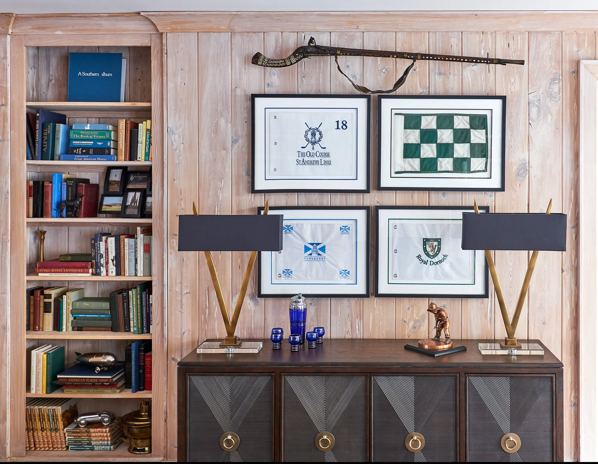 A built-in wooden bookcase is filled with stacked books and decor. On the adjacent paneled wall, four framed golf flags hang above a dark wood console.