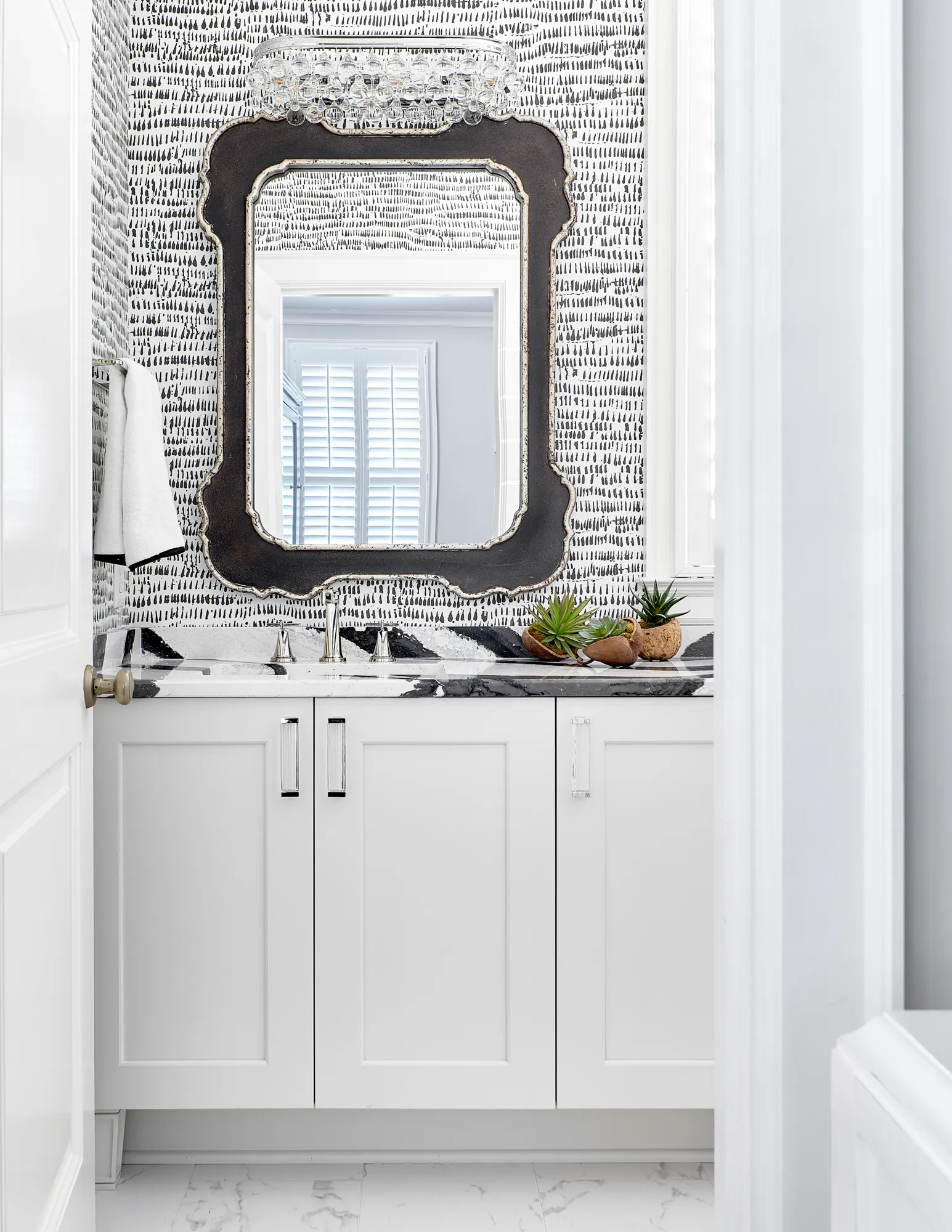 A powder room with high-contrast black and white decor. A white vanity and marble counter sit against bold patterned wallpaper, topped by an ornate mirror.