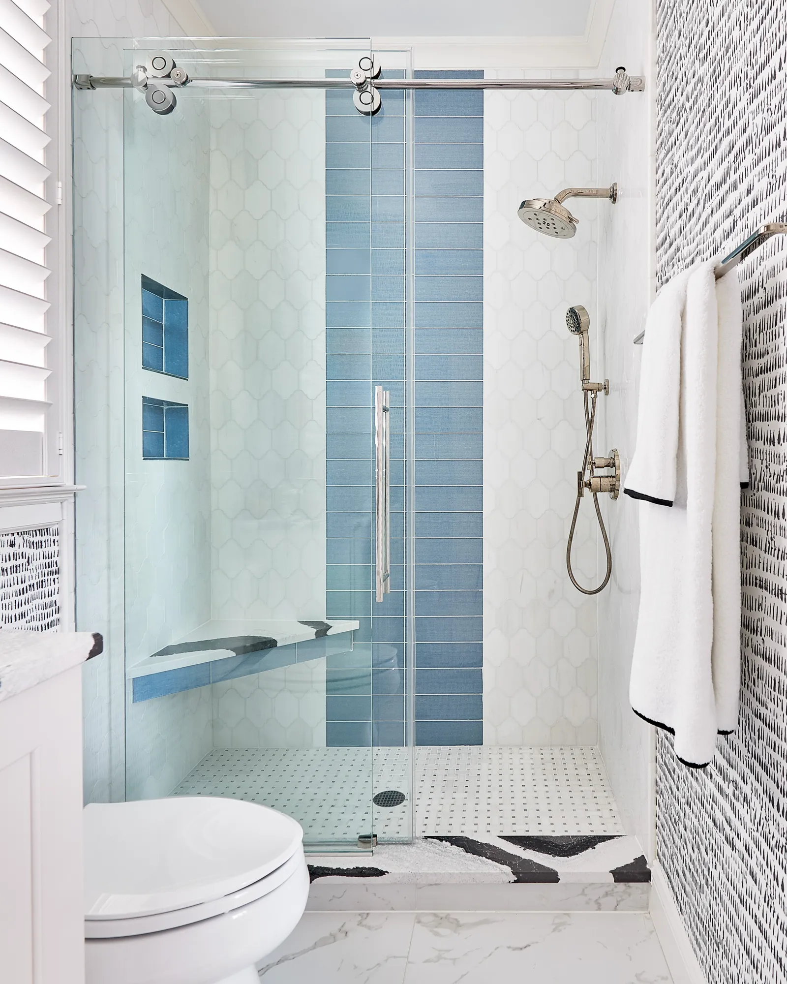 A contemporary walk-in shower with glass doors features white honeycomb tiles, a vertical band of blue subway tiles, a built-in bench, and chrome fixtures.