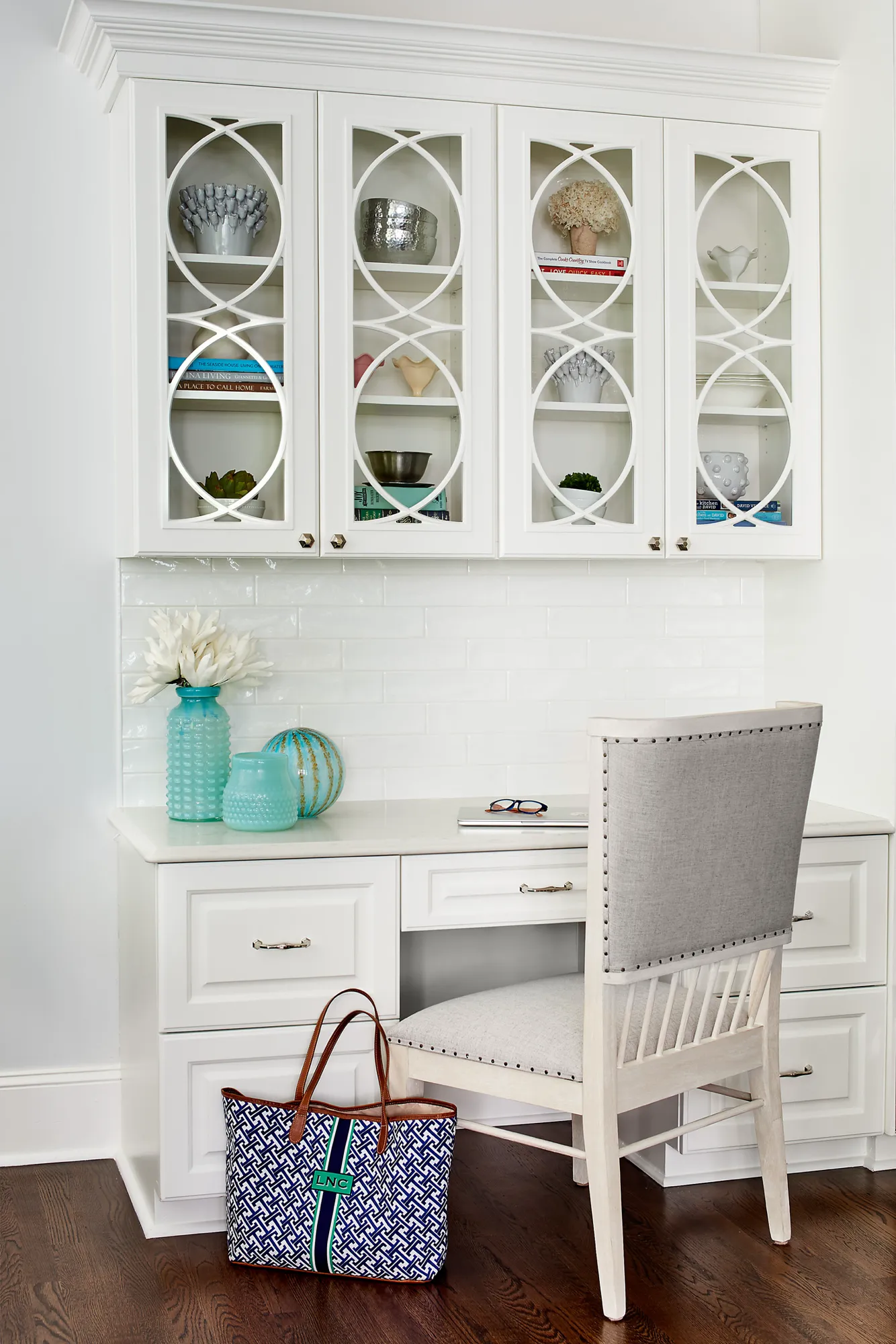 A clean white built-in home office desk features decorative upper cabinets with lattice glass doors and a subway tile backsplash. A gray upholstered chair is pulled to the desk.