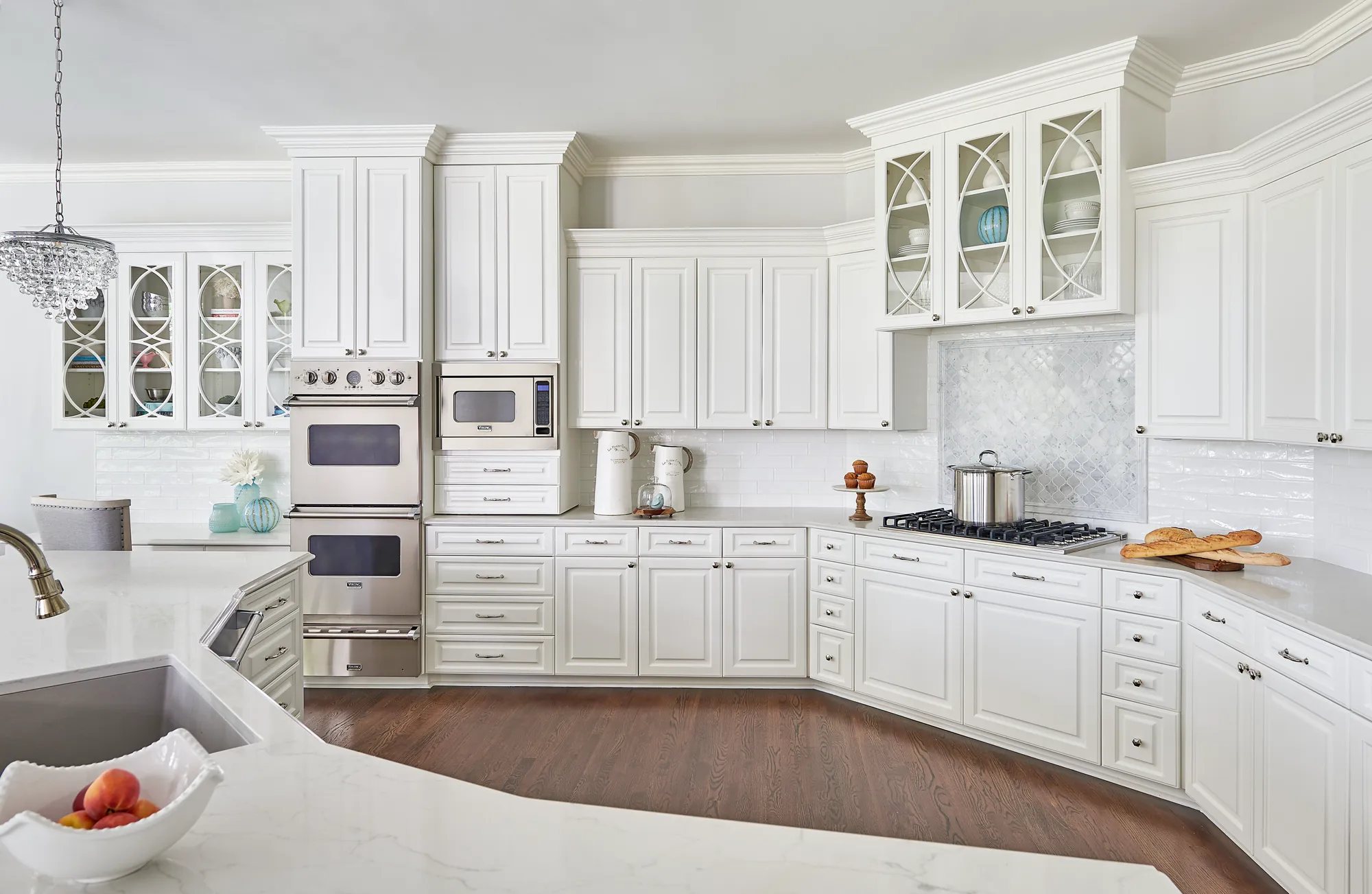 An upscale white kitchen with detailed glass-front upper cabinets, marble countertops, stainless steel appliances, and dark wood flooring.