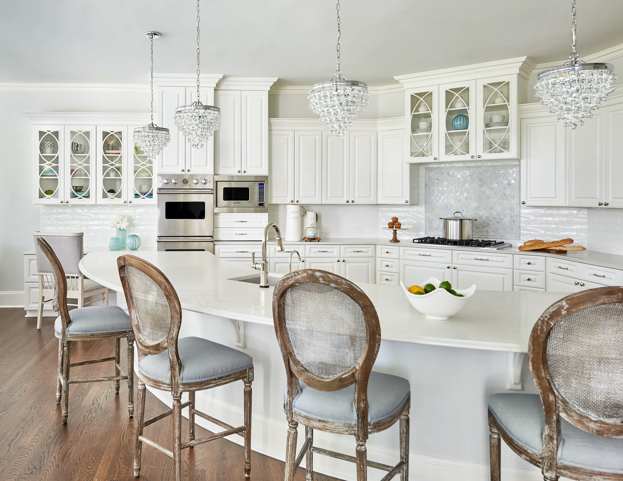 A luxury white kitchen features crystal chandeliers above a large island and four distressed wood stools. White cabinets frame stainless steel appliances and a decorative tile backsplash.