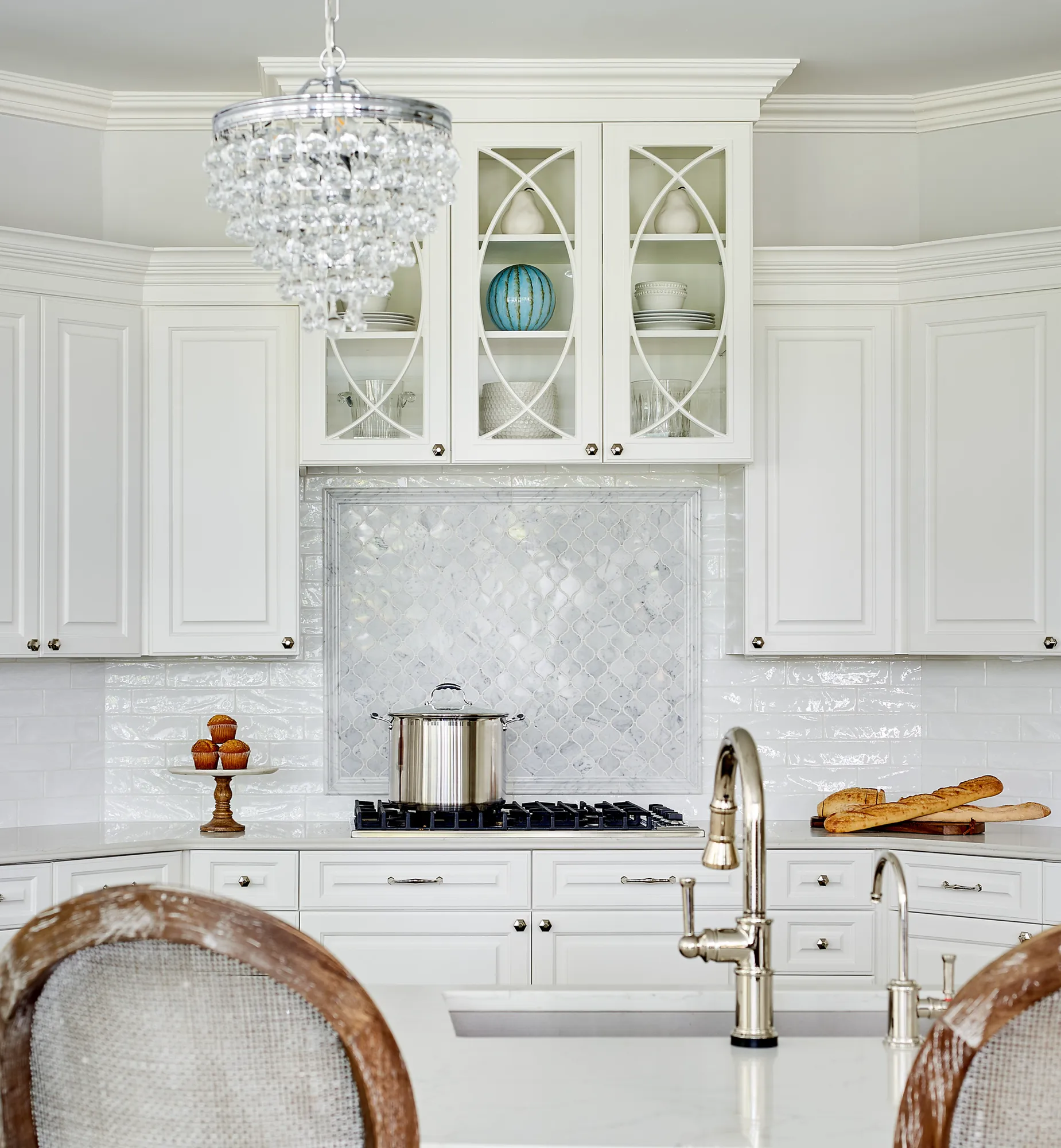 A high-end white kitchen featuring elegant custom cabinets, a crystal chandelier, and a stove with a polished steel pot. The backsplash uses glossy subway tiles and a marble mosaic accent.
