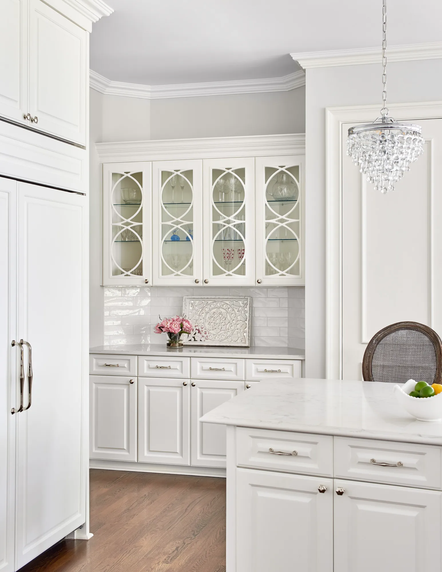 An elegant white kitchen with an integrated refrigerator and a custom hutch featuring decorative glass doors, marble counters, and a small crystal chandelier.