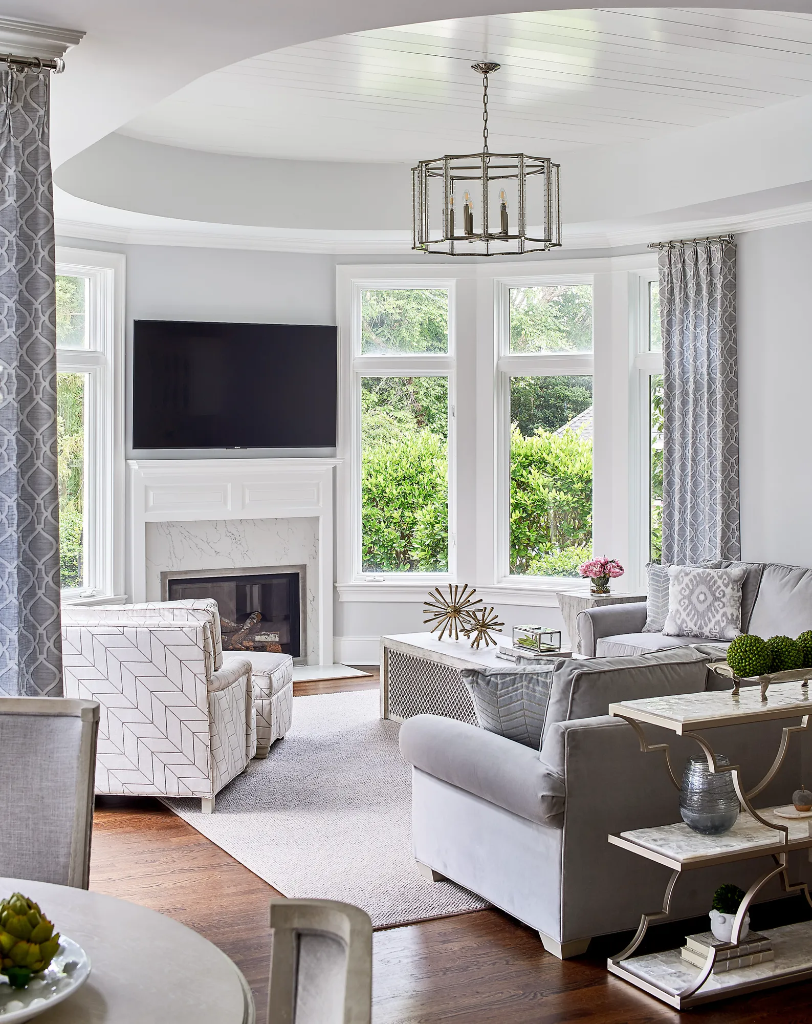 A bright, elegant living room featuring a bay window overlooking lush greenery. The room has gray upholstered furniture, patterned accent chairs, a white fireplace with a mounted TV, and a metallic chandelier.