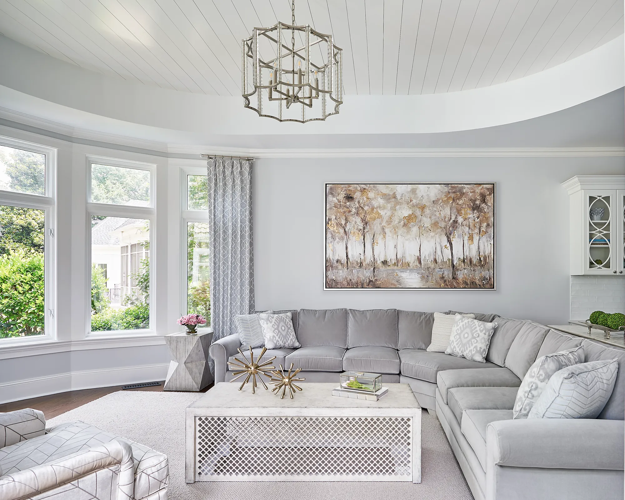 An elegant living room featuring a large gray velvet sectional sofa, a white coffee table, and an abstract forest painting. A curved paneled ceiling highlights the bright bay window area.