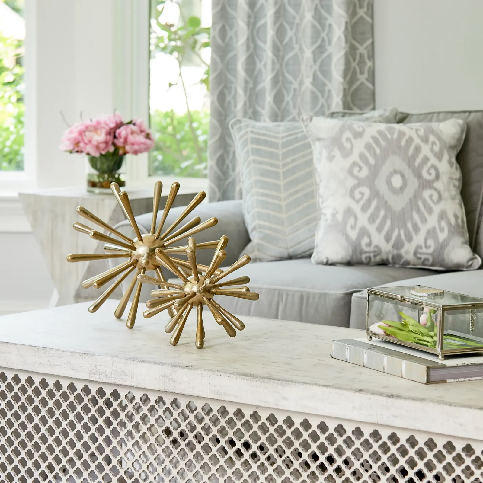 A gold starburst sculpture sits on a distressed white coffee table in a bright living room. In the background is a gray sofa with patterned pillows.