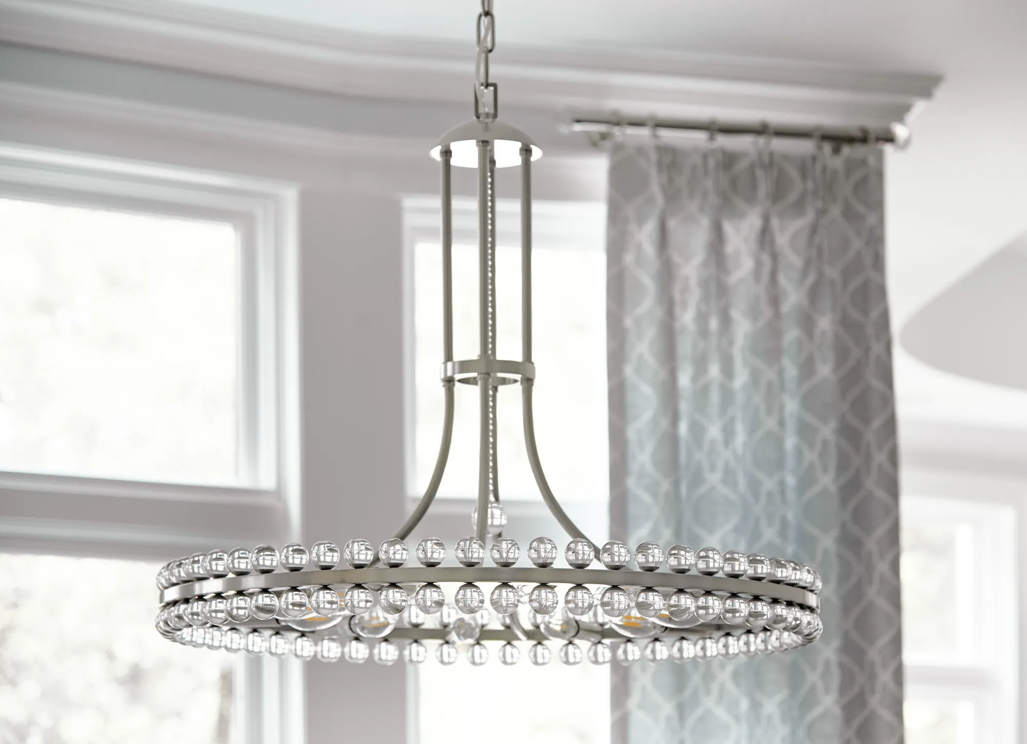 An elegant brushed nickel chandelier hangs from the ceiling, featuring a wide circular frame lined with clear glass spheres in a room with bright windows.