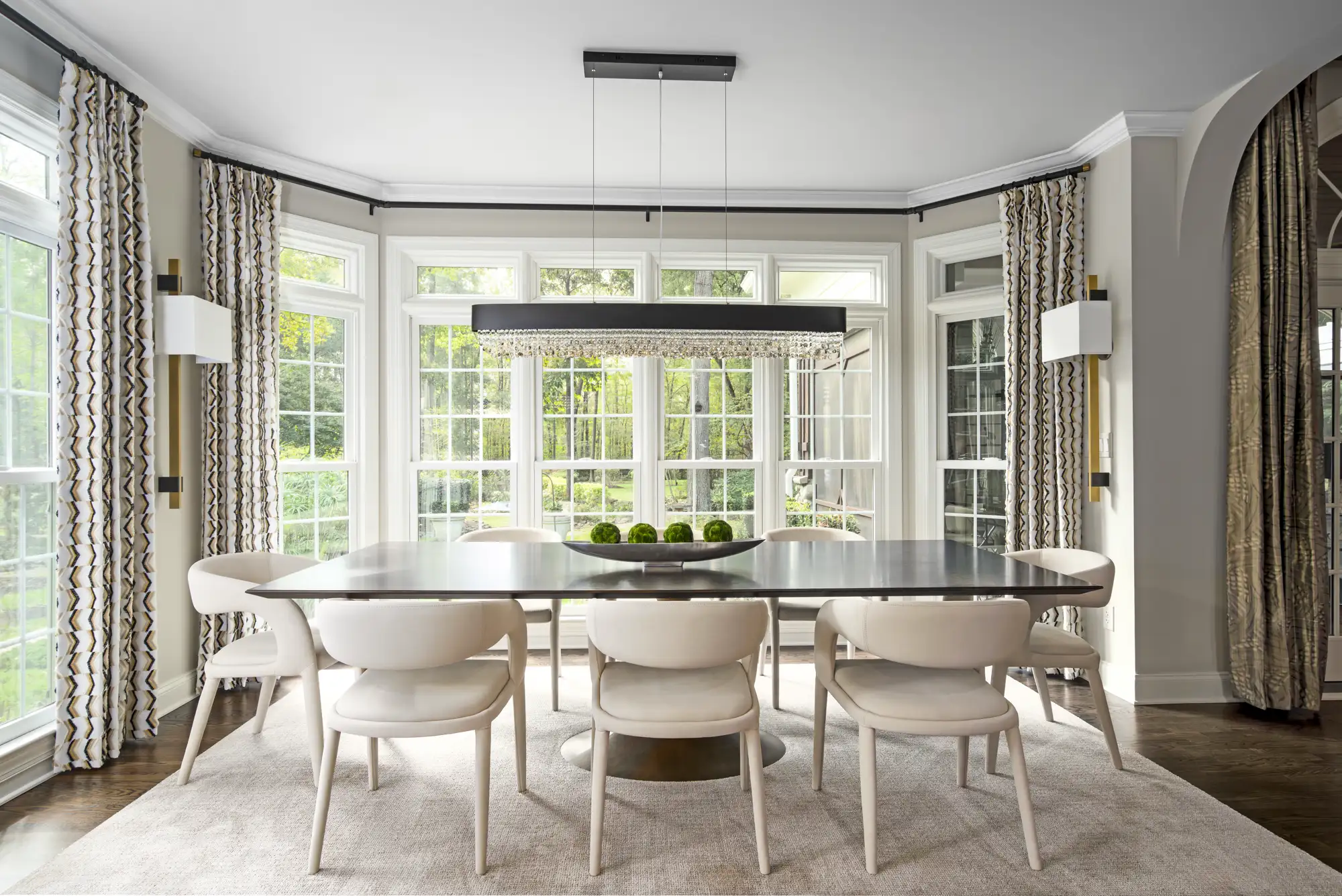 An elegant dining room featuring a dark rectangular table and modern cream chairs, centered under a linear crystal chandelier and framed by bay windows.