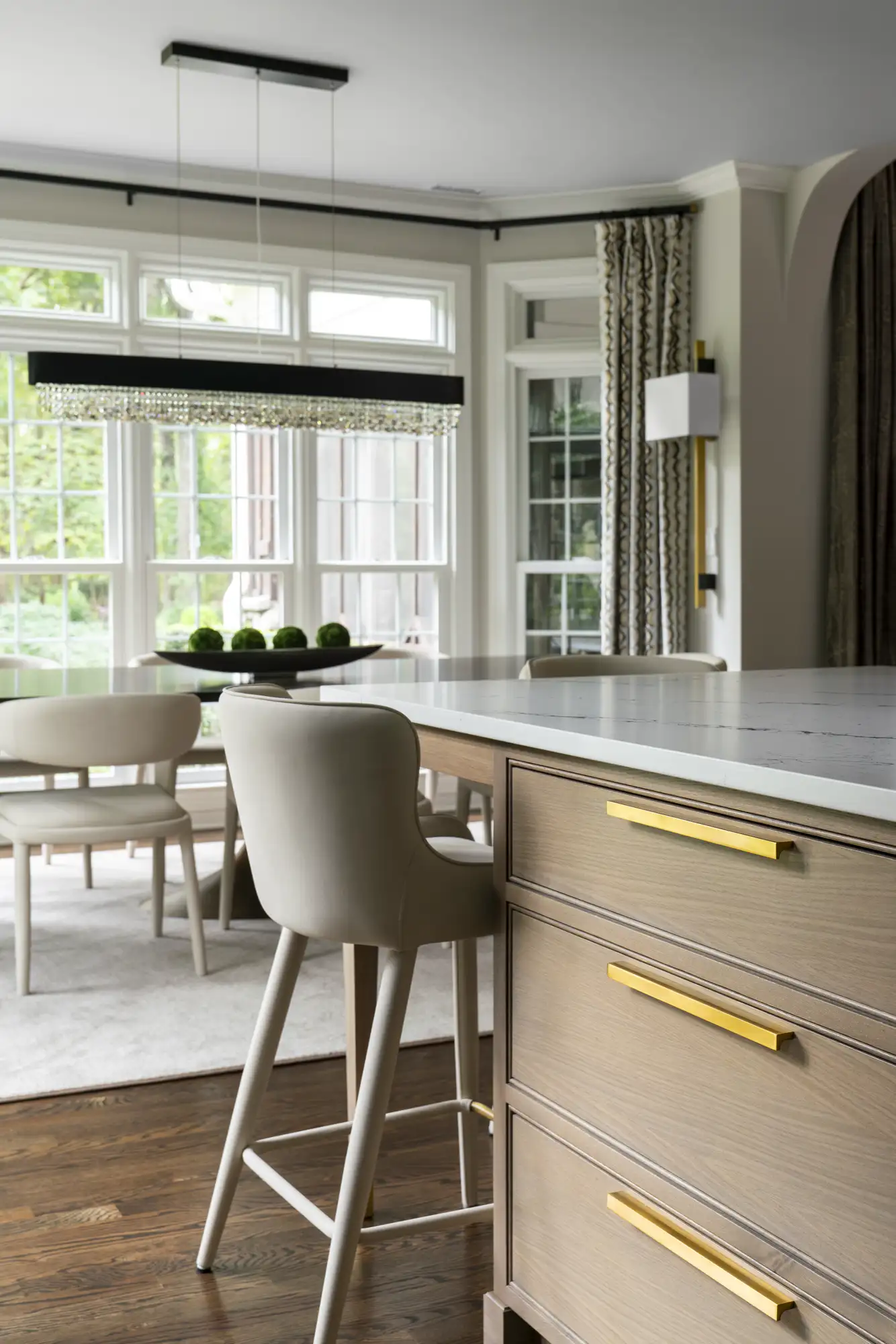 A luxurious kitchen island with light wood cabinetry and gold bar pulls stands near a dining area illuminated by large windows and a rectangular crystal chandelier.