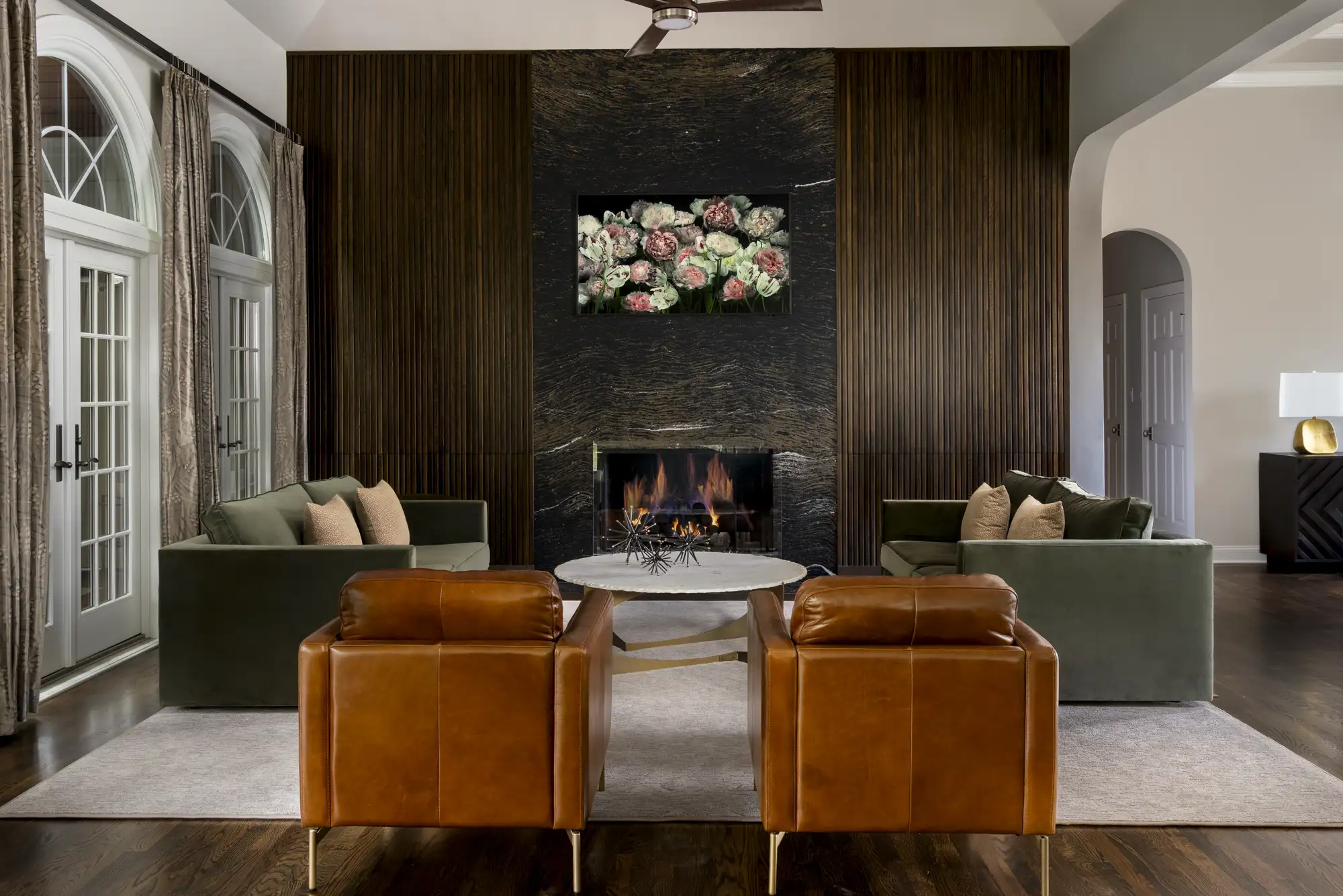 A modern living room with dark wood floors and high ceilings. Tan leather chairs and olive green sofas frame a marble table in front of a fireplace wall with dark wood slats.