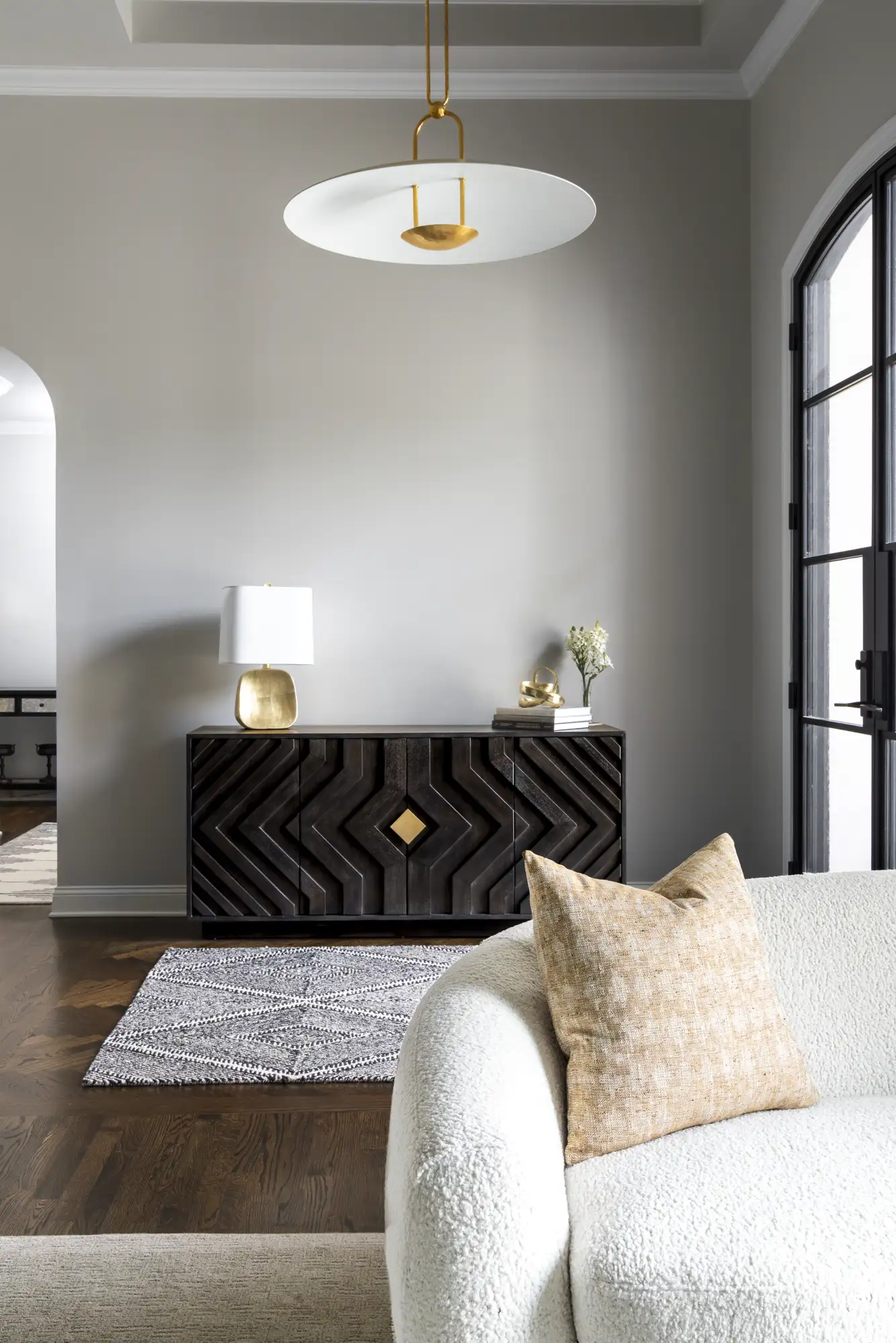 A dark wood credenza with a geometric chevron pattern sits against a gray wall, illuminated by a white and gold pendant light. A gold lamp and flowers decorate the top.