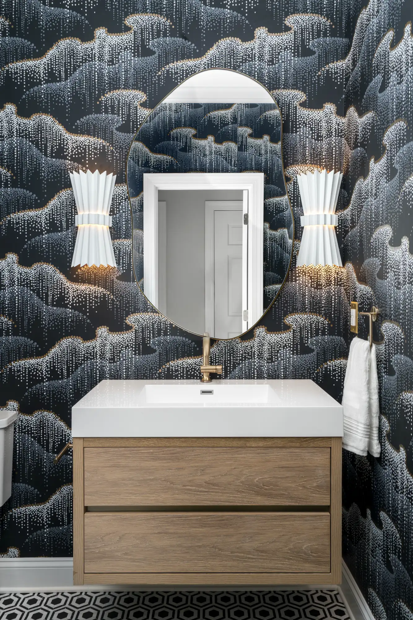 A modern powder room vanity area with dark, dotted wallpaper. It features a light wood floating cabinet, a gold-framed oval mirror, and two white pleated sconces.