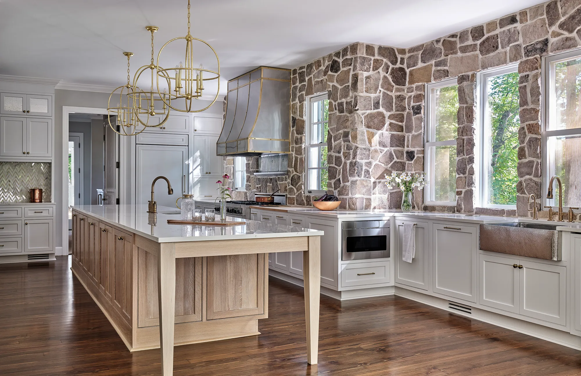 A luxury kitchen with a rustic stone wall contrasting with light cabinets and a wood island. Custom gold chandeliers and a large metal hood define the space.