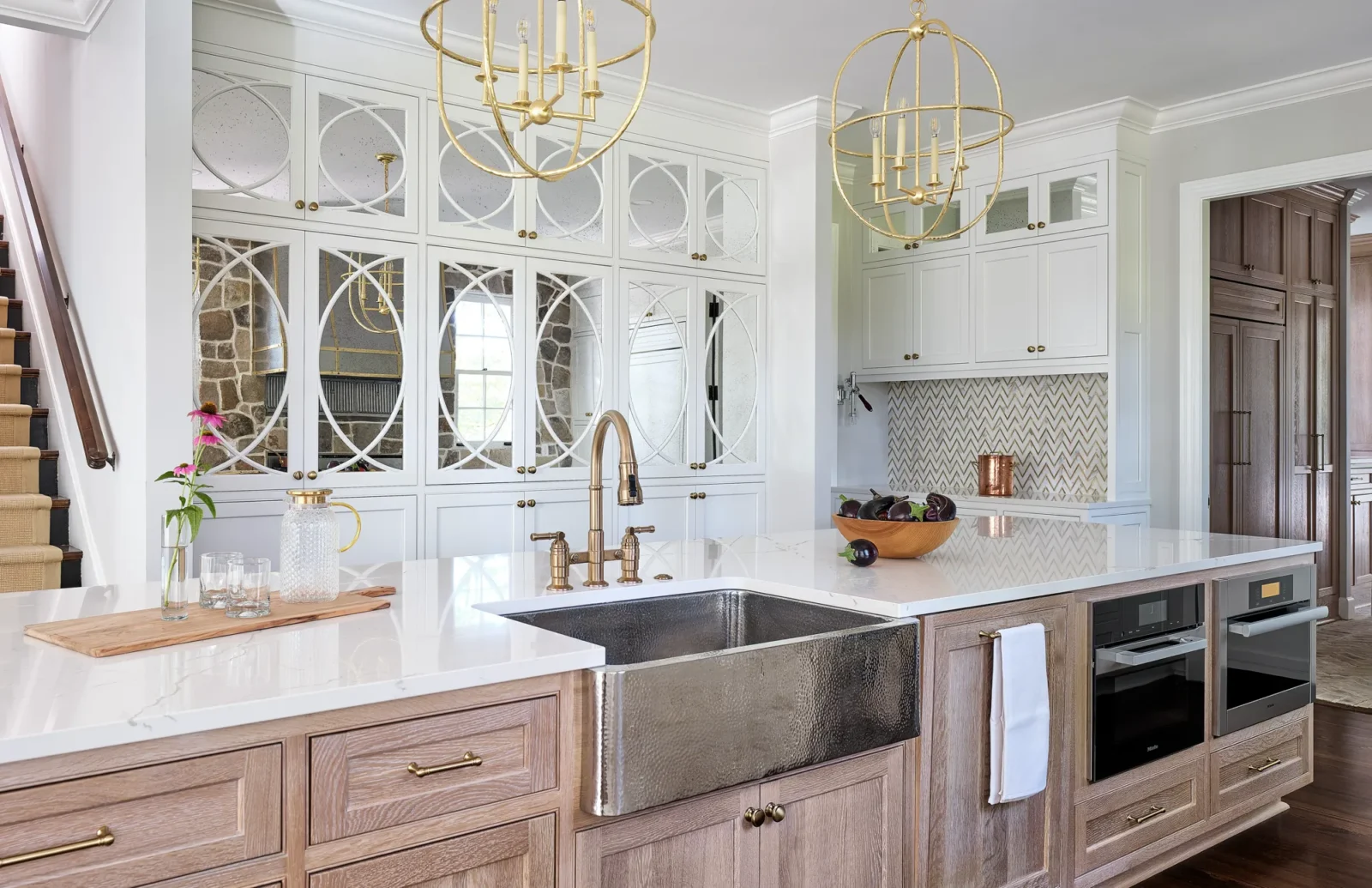 A luxurious kitchen with natural wood island cabinets, a farmhouse sink, and brass fixtures, contrasting with white wall cabinets that feature mirrored, geometric door panels.