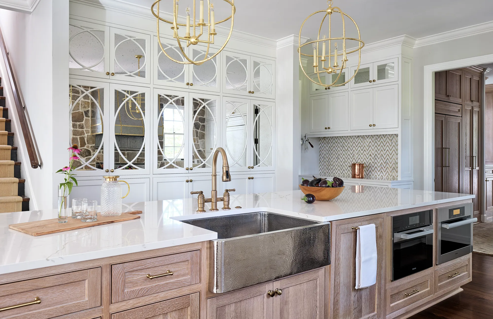 An elegant kitchen island featuring a hammered metal sink, brass faucet, and white countertop. The back wall has white cabinetry with decorative mirrored panels and brass globe light fixtures.