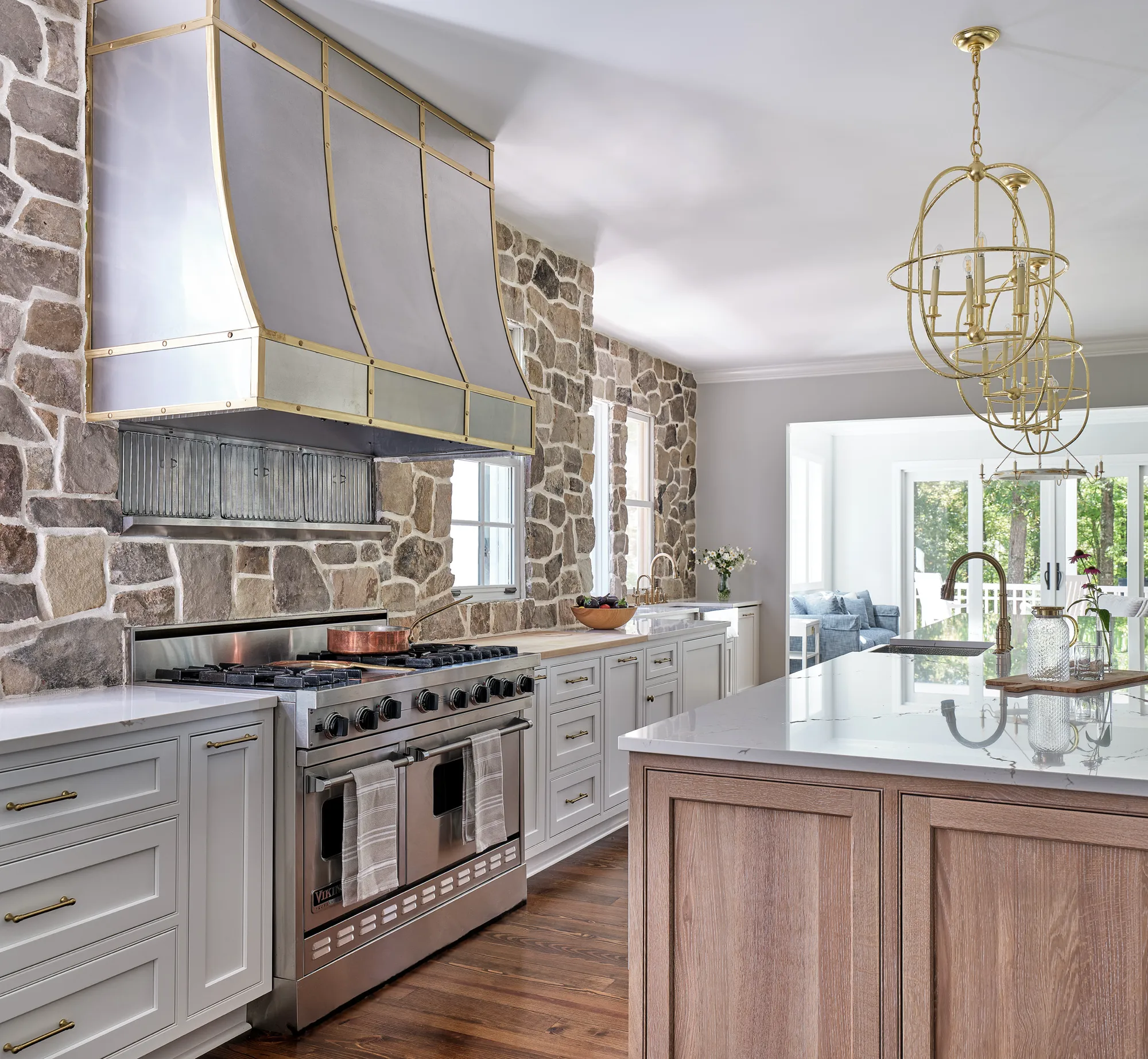 A luxury kitchen featuring a large stainless steel range and custom brass-trimmed metal hood against a rustic stone accent wall. Light gray cabinets and a natural wood island blend the styles.
