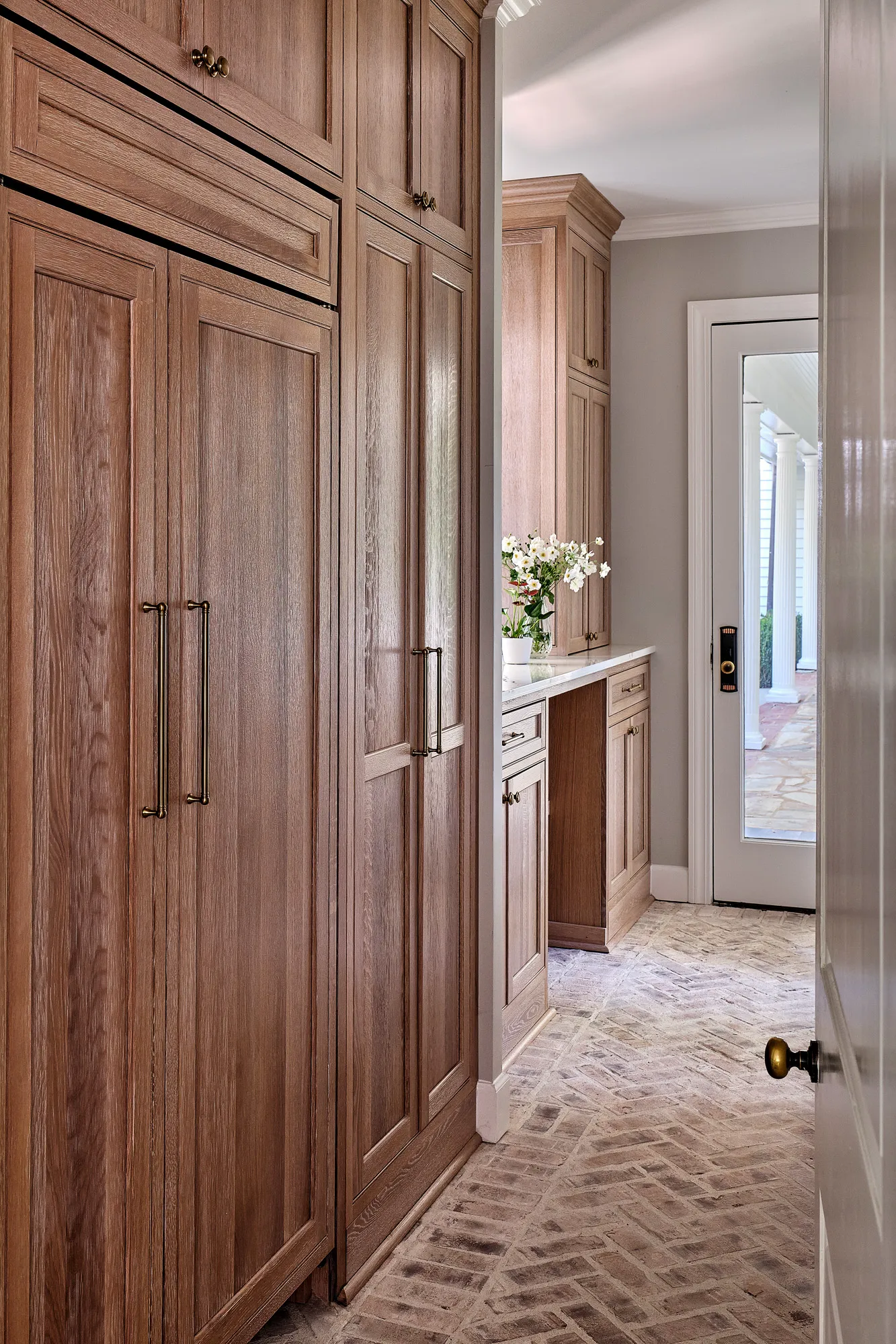 Custom wood cabinetry lines a hallway with a white marble counter and a brick herringbone floor. A glass door leads to a columned outdoor porch.