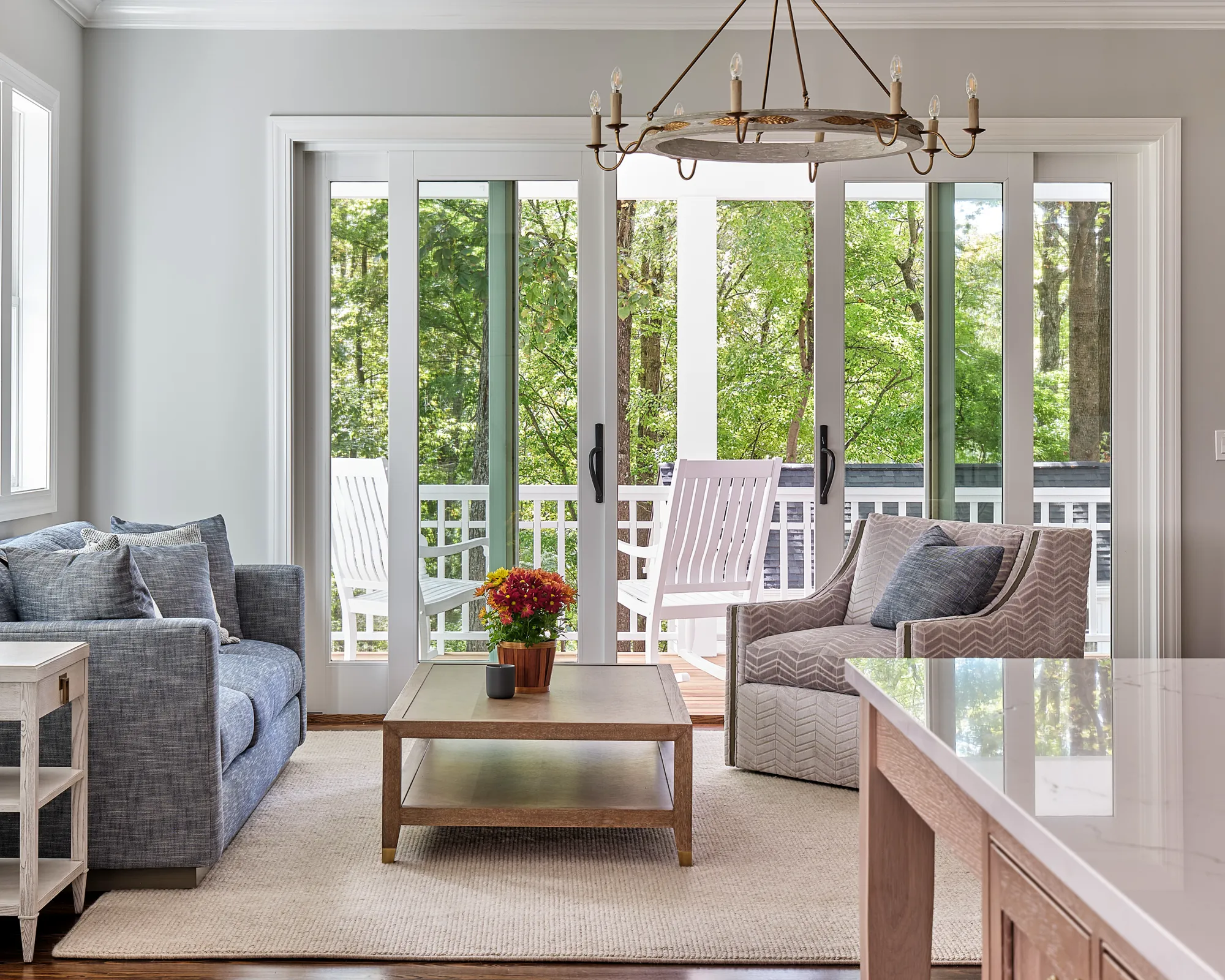 A bright living space featuring a blue sofa and patterned armchair, centered around a coffee table. Sliding glass doors open to a deck and reveal a tranquil view of lush green trees.