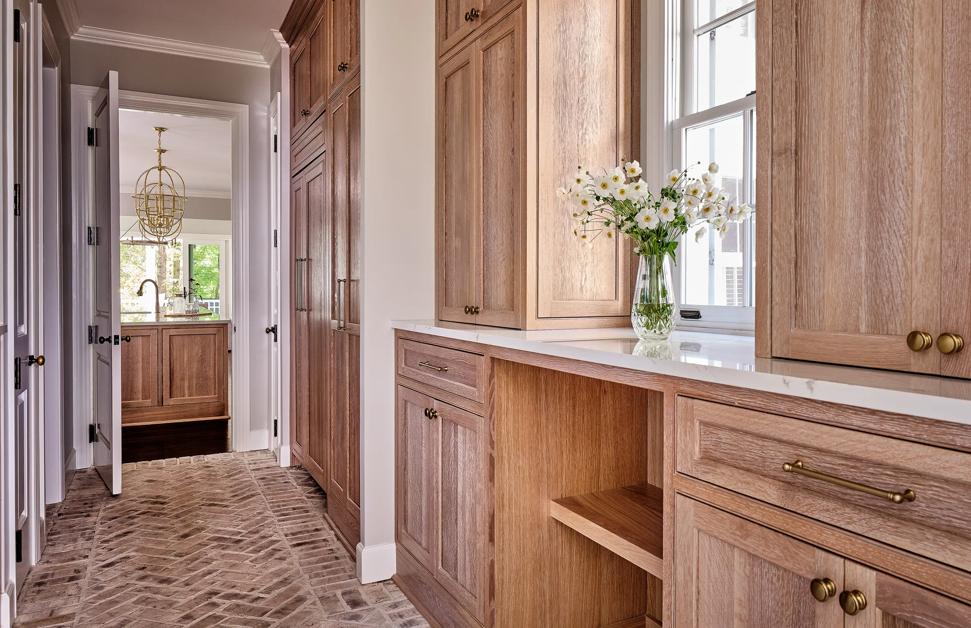 A corridor featuring herringbone brick flooring and custom light wood cabinetry with brass hardware. White flowers sit on a counter near a window.