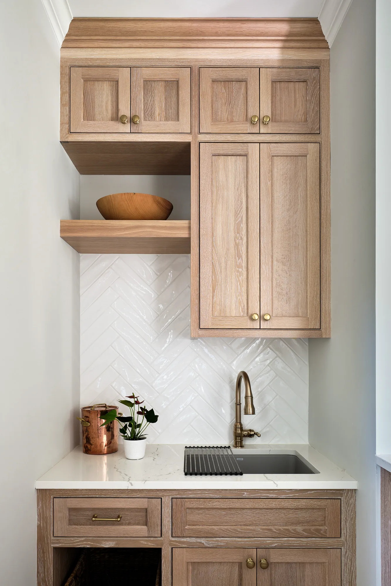 A built-in cabinet unit with light wood doors, a white quartz countertop, and a sink with a brass faucet. The backsplash features white herringbone tiles.