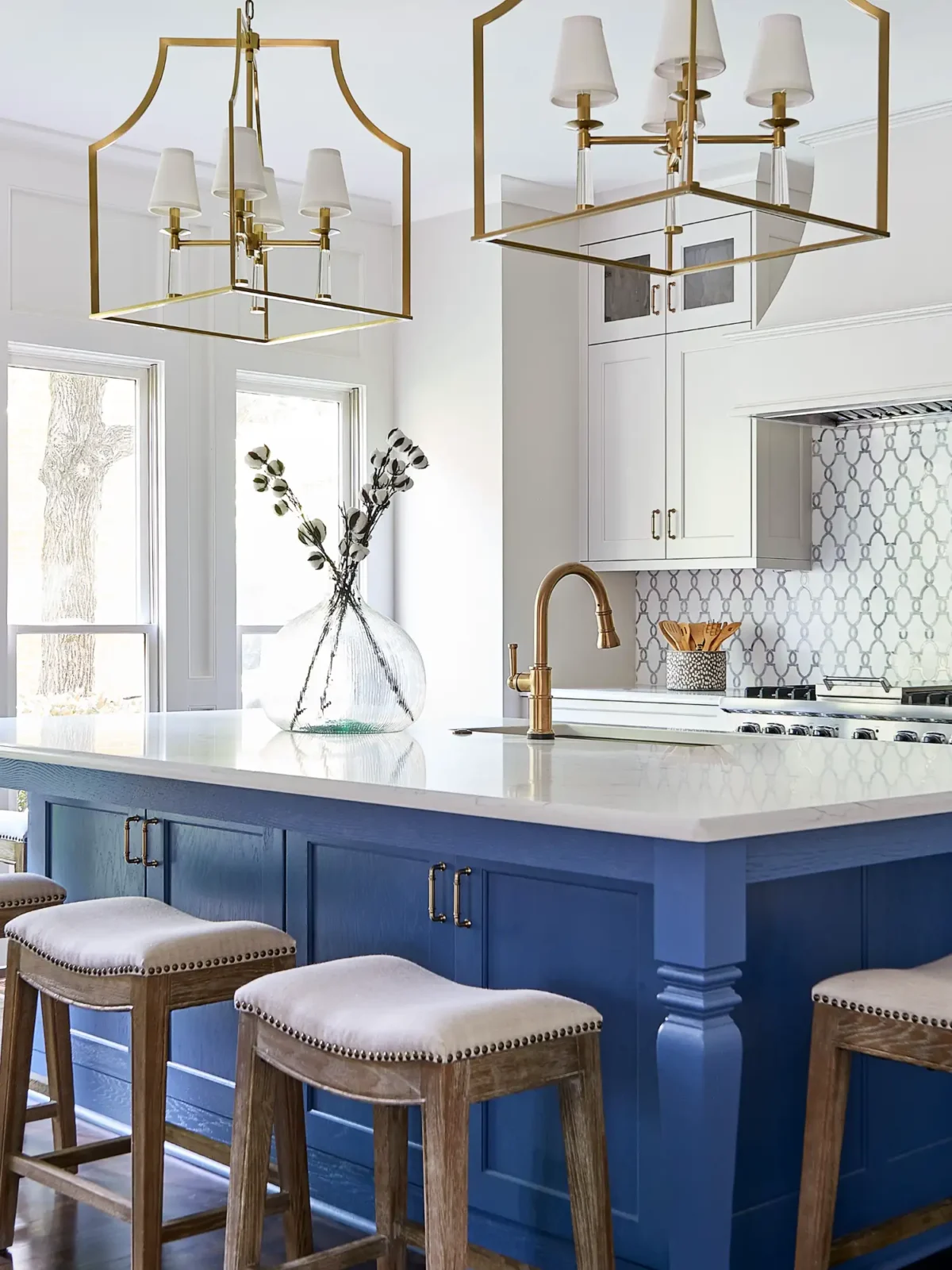A modern, bright kitchen has a large royal blue island with white quartz counters. Two brass lantern pendants hang above, complementing the brass faucet.