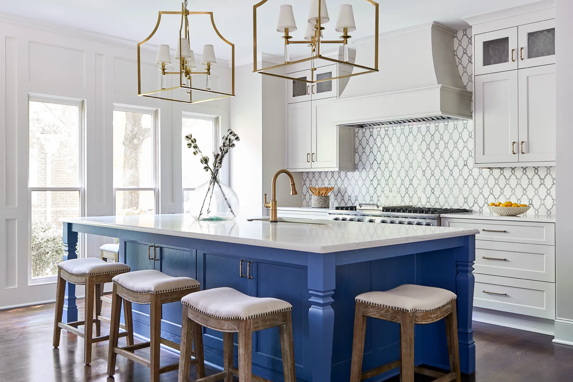 A bright kitchen features a large blue island with four stools, contrasted by white cabinets and a decorative patterned tile backsplash. Two brass lantern lights hang overhead.