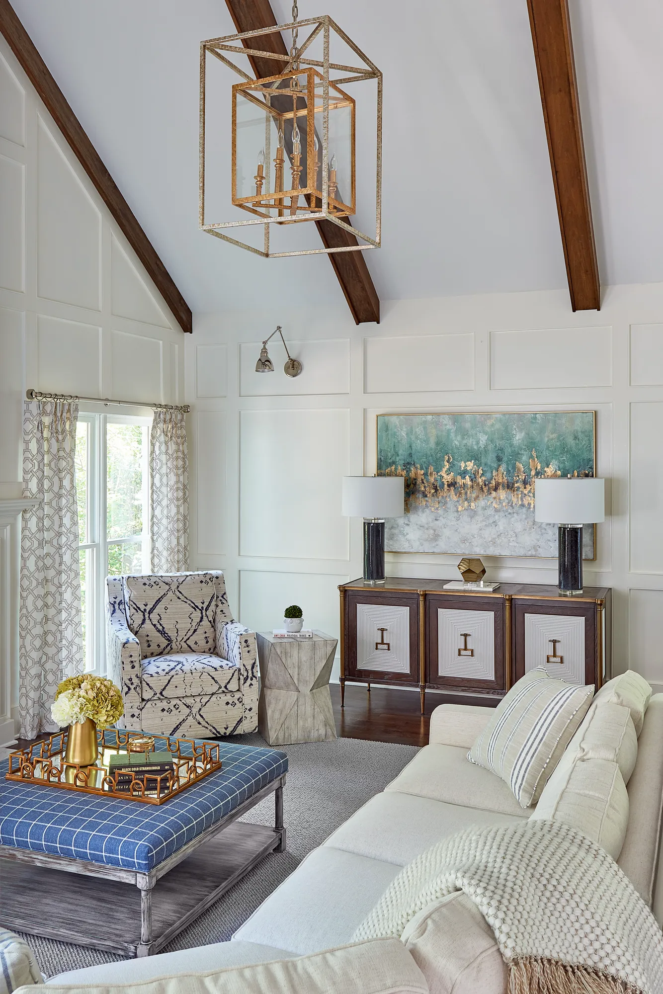 An elegant living room features a vaulted ceiling with wood beams and white paneled walls. A gold cage chandelier hangs over a plaid ottoman and a comfortable white sofa.