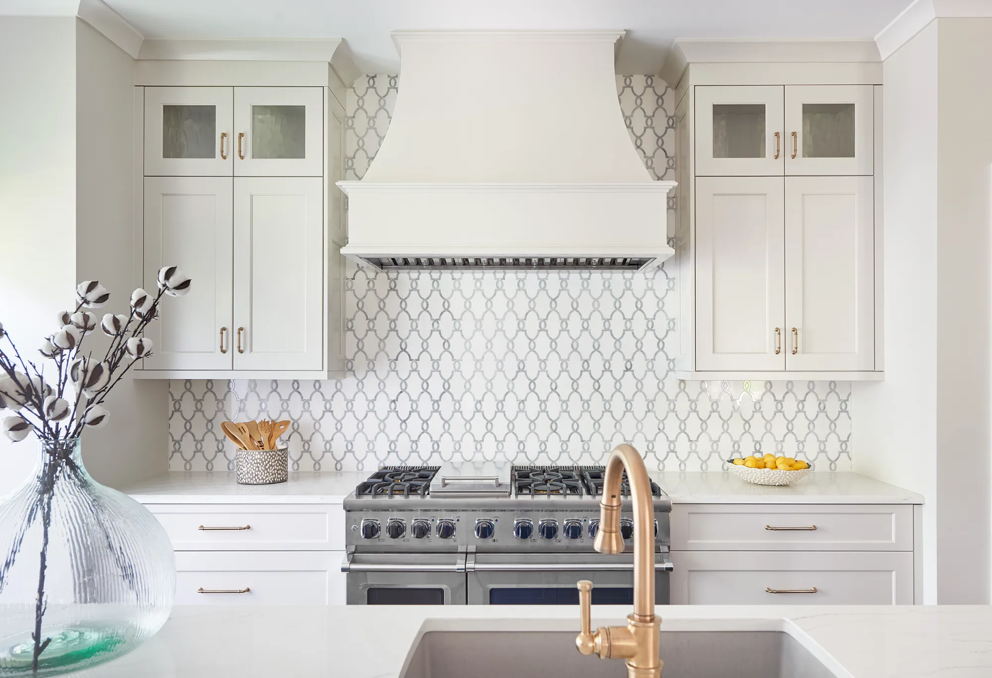 A luxurious white kitchen with shaker cabinets, a brass faucet, and a stainless steel range set against an ornate gray geometric tile backsplash.