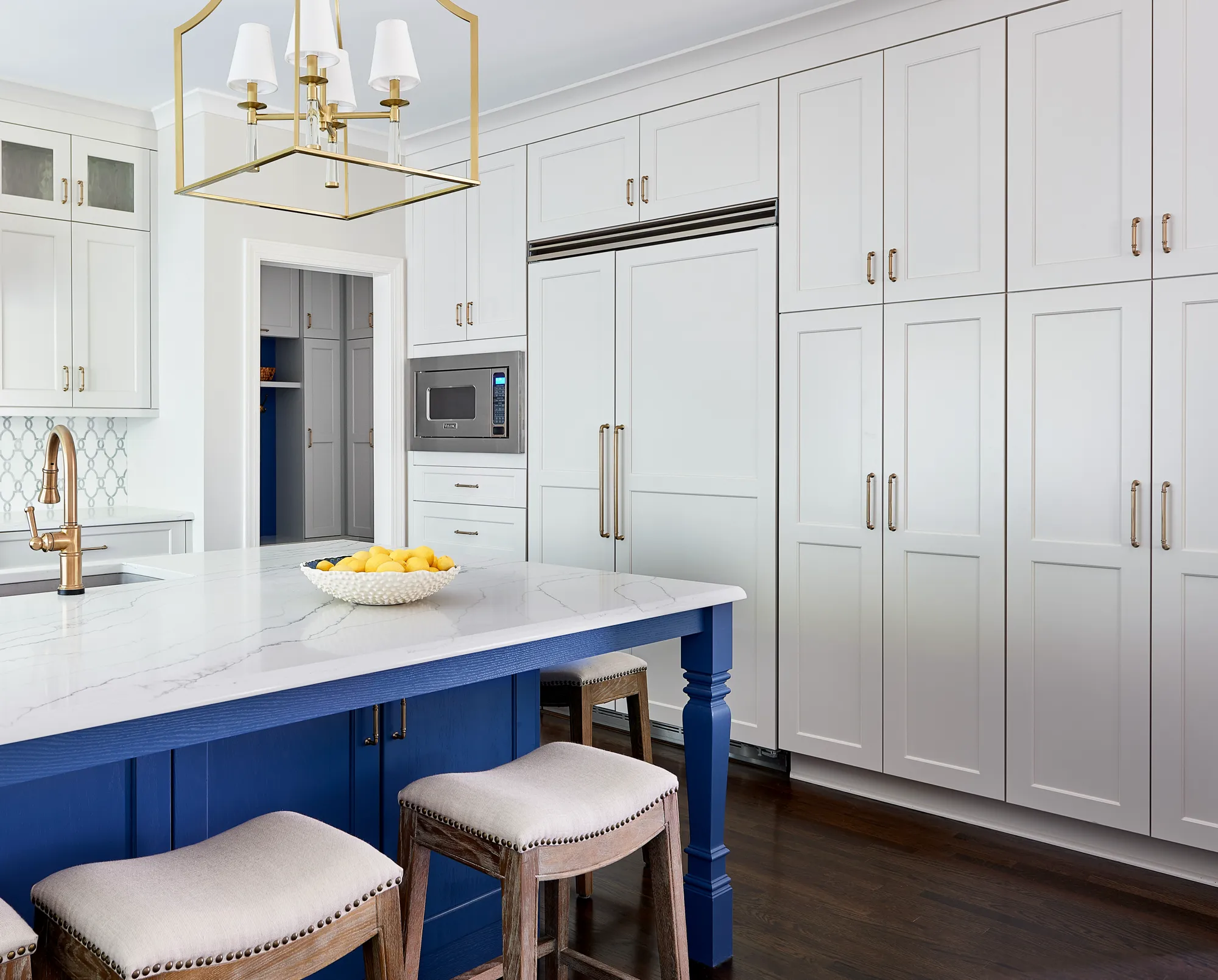 A modern, custom kitchen featuring extensive white cabinetry and brass hardware. The bright blue island is topped with marble, contrasting with the dark wood floor.
