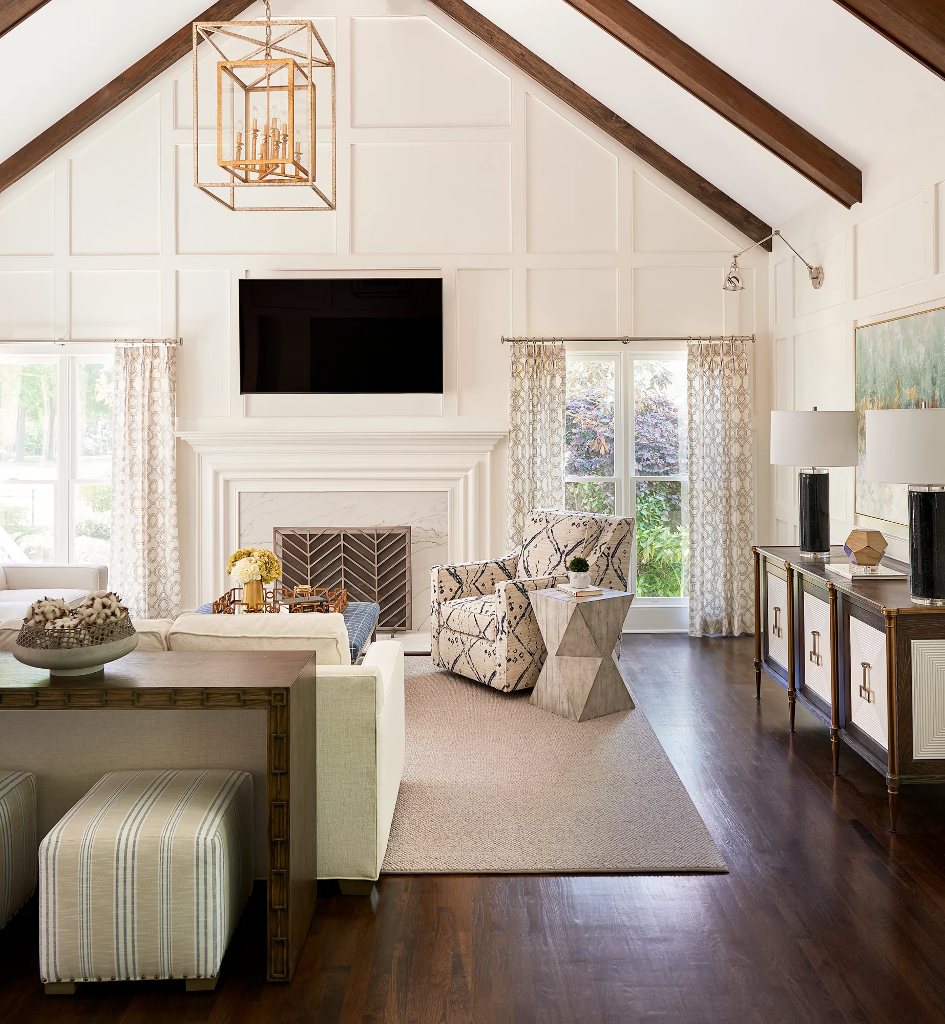 A brightly lit, elegant living room features a vaulted ceiling with dark wood beams. White paneled walls surround a fireplace and a mounted TV above.