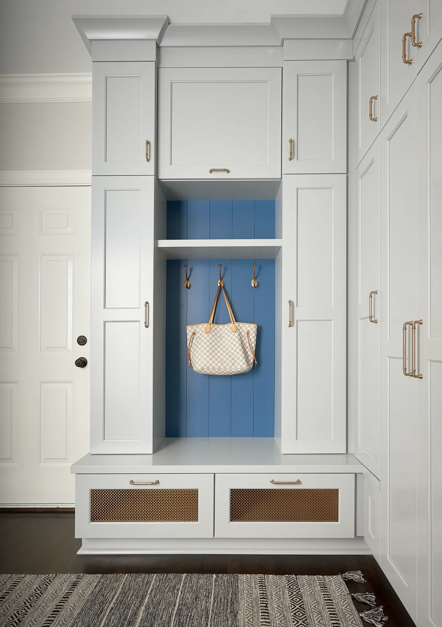 A white built-in mudroom unit has a bench, upper storage, and drawers with gold mesh inserts. A patterned tote hangs in the central blue cubby.