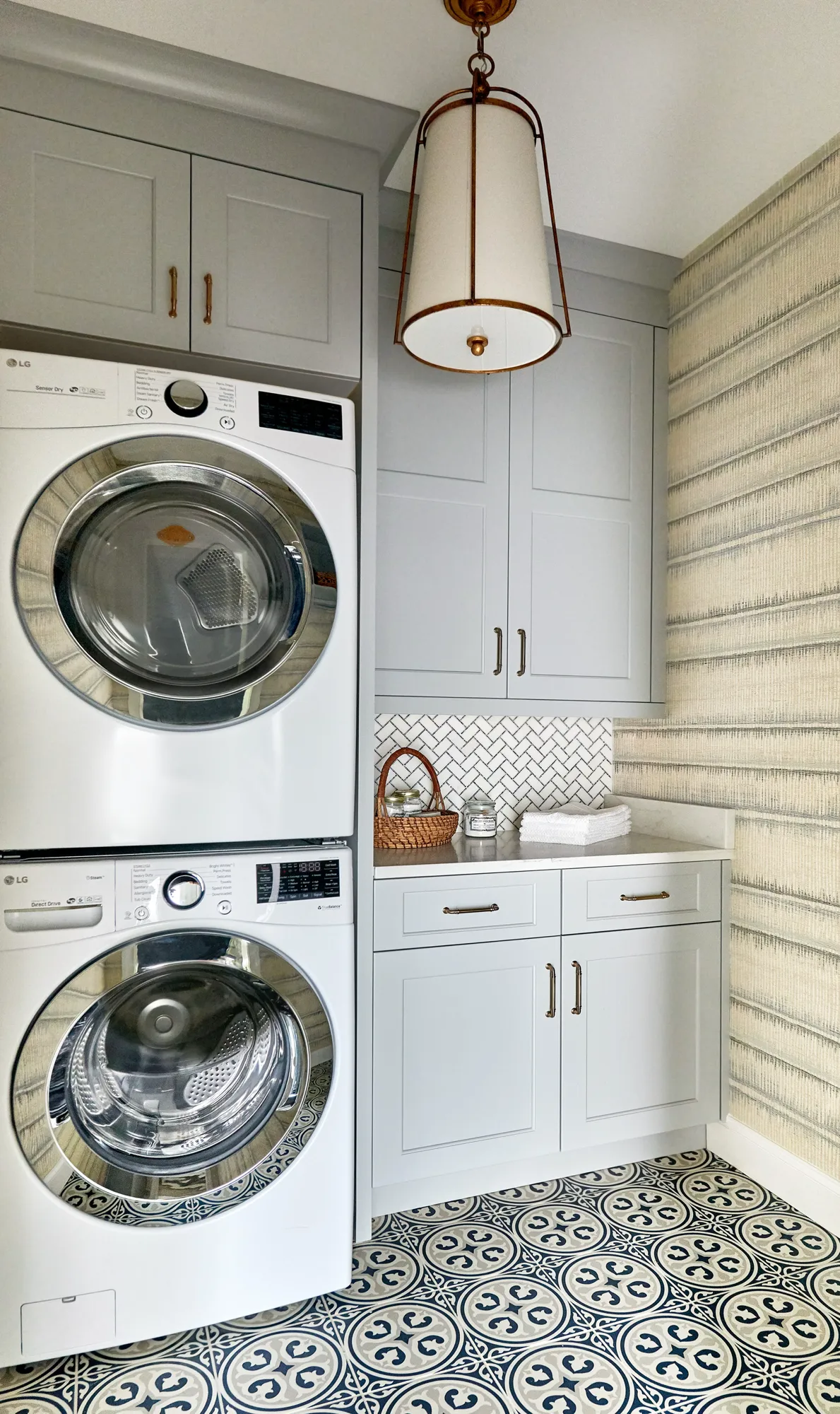 A stylish laundry room with stacked washer and dryer units next to light gray cabinetry with brass pulls and a white herringbone backsplash. The floor has a bold patterned tile.