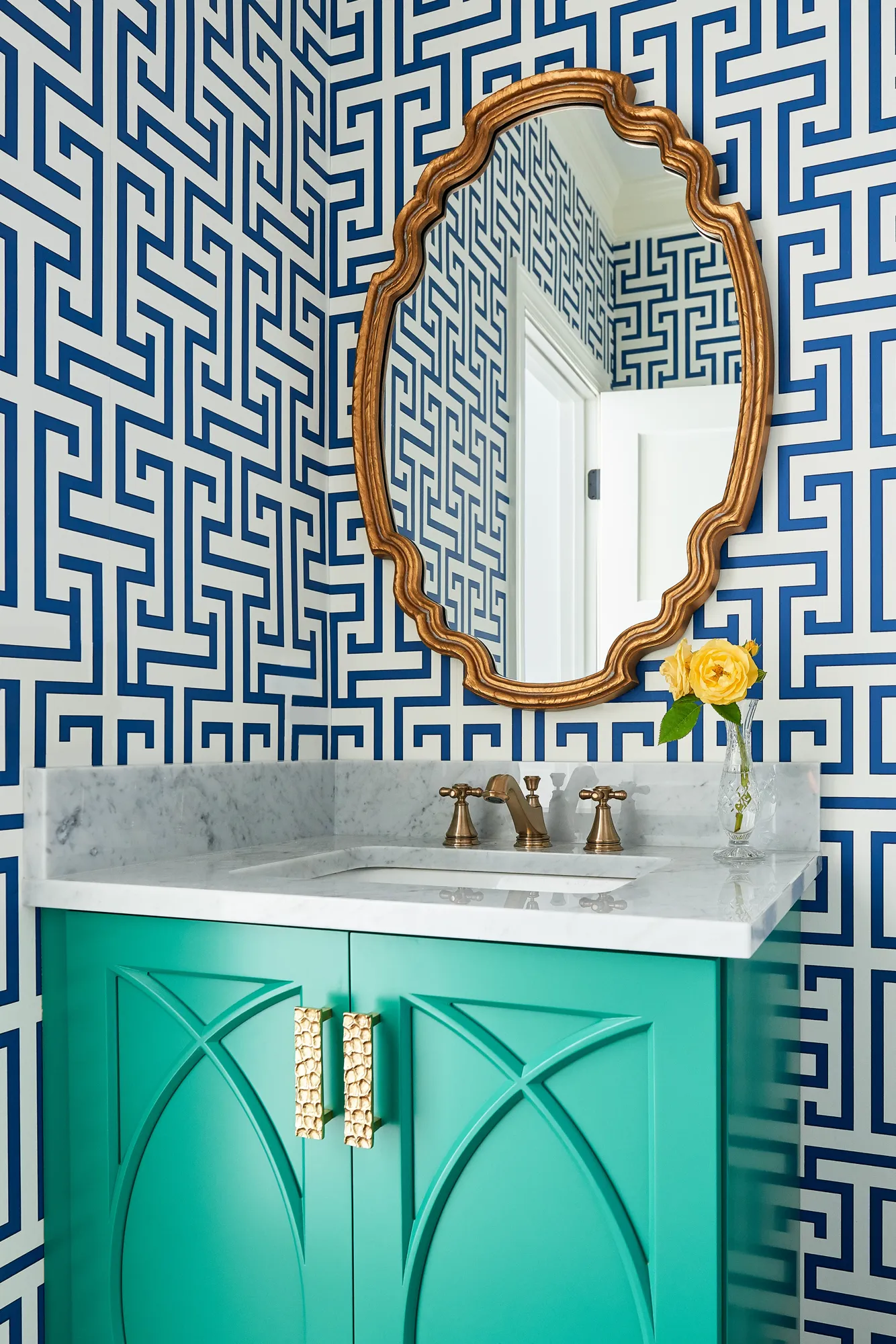 A striking powder room with blue Greek key wallpaper and a teal vanity with gold hardware. An ornate gold mirror hangs above the marble counter.