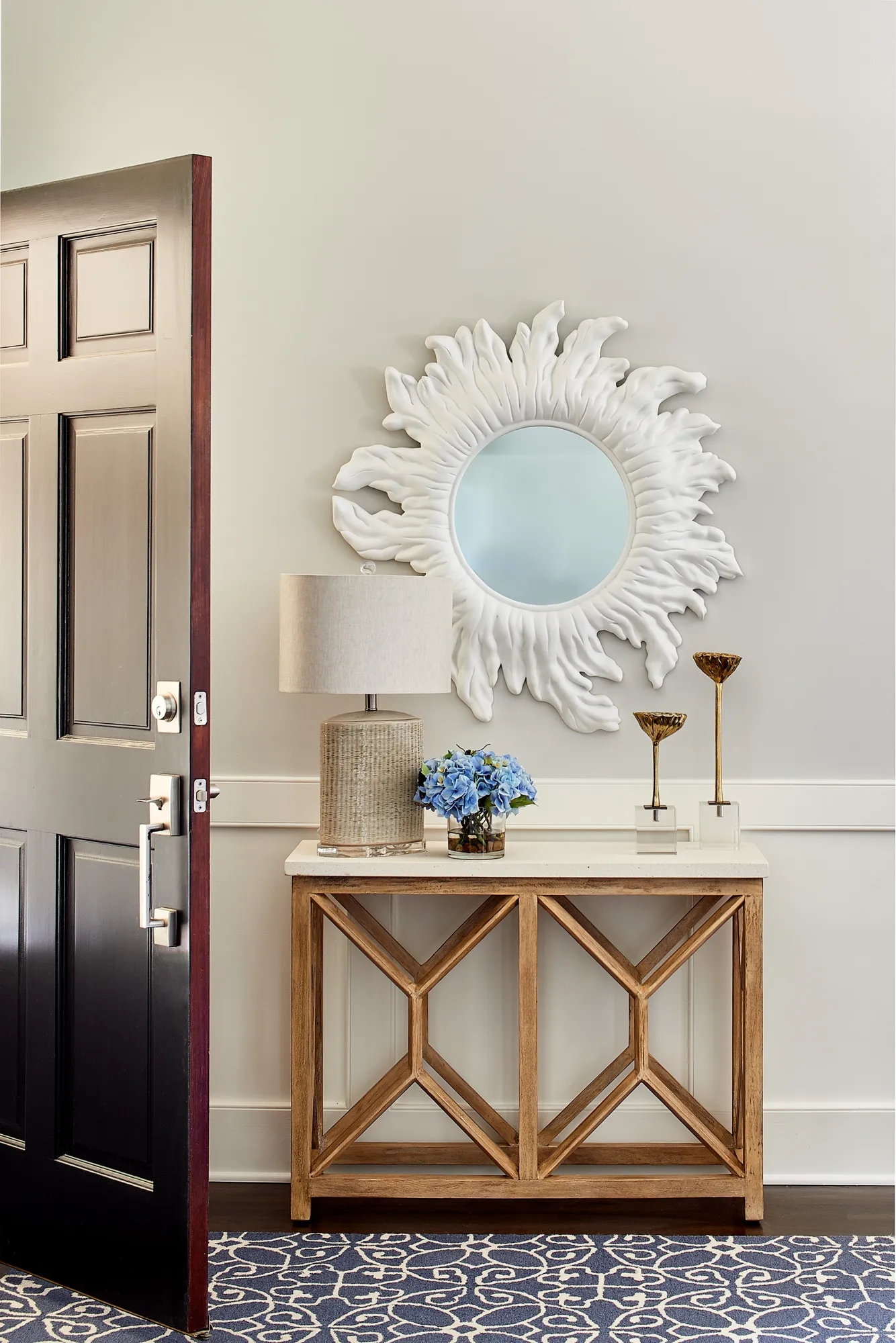 An entryway vignette featuring a wooden console table with a white marble top, holding a lamp and blue hydrangeas, set below a white sunburst mirror.