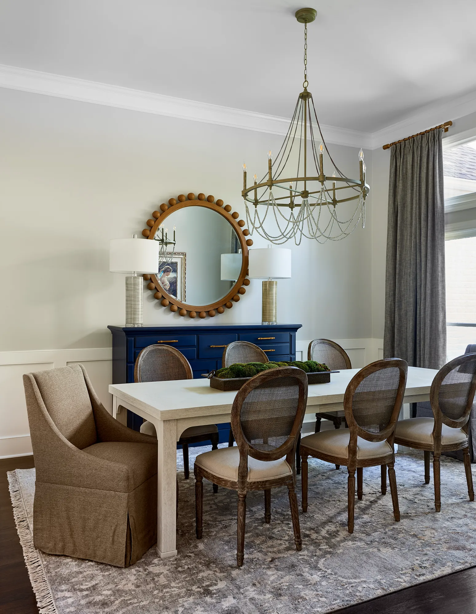 A modern dining room with a white table, cane-back chairs, and a large navy blue credenza. A circular wood-beaded mirror and antique brass chandelier add detail.