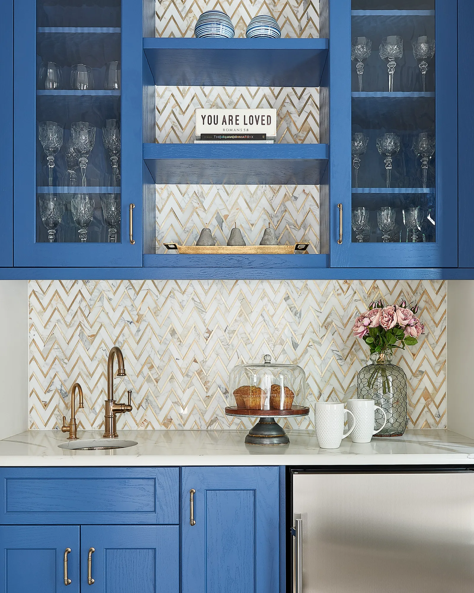 A wet bar featuring cobalt blue cabinets, a white and gold chevron backsplash, and a bronze faucet. Glassware and pink roses decorate the marble counter.