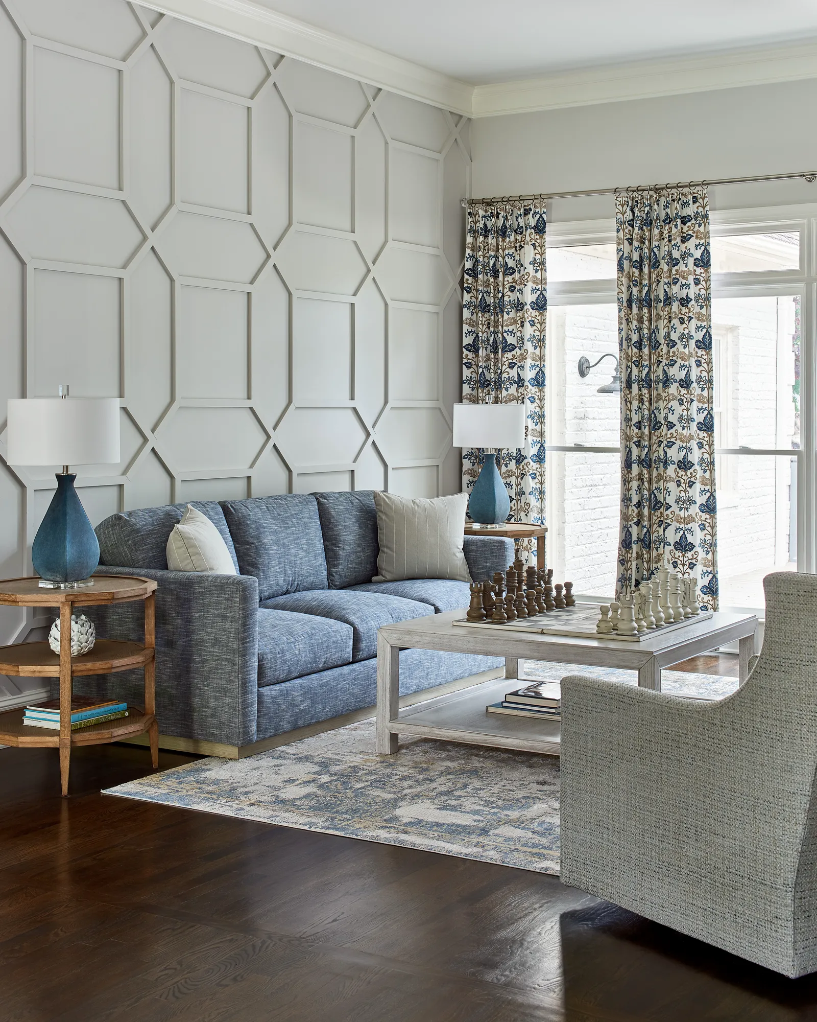 A stylish living room with a blue sofa facing a coffee table holding a full chess set. The room features dark wood floors and geometric wall paneling.