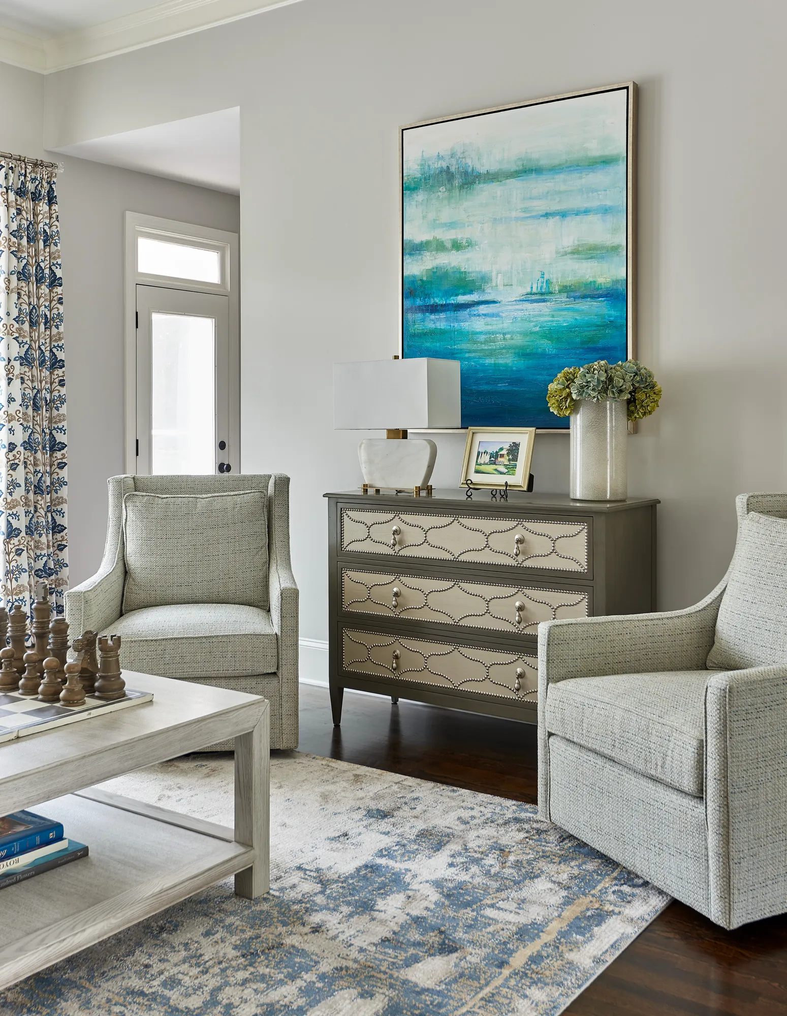 An elegantly decorated sitting area featuring two light gray upholstered chairs framing a studded gray chest under a large abstract teal painting.