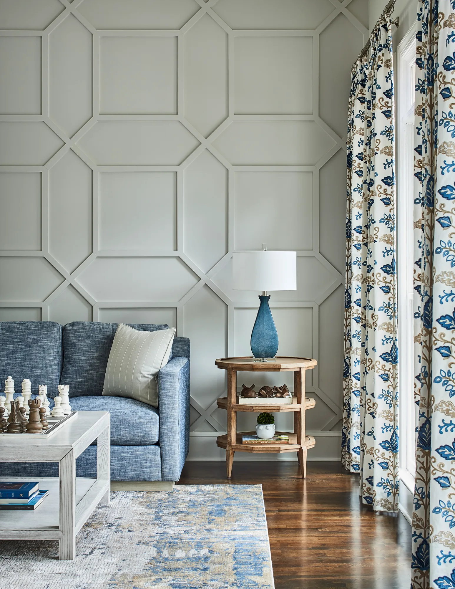 An elegant living room features a blue sofa next to a geometric accent wall. Blue floral curtains frame a window, complementing the rug and table lamp.