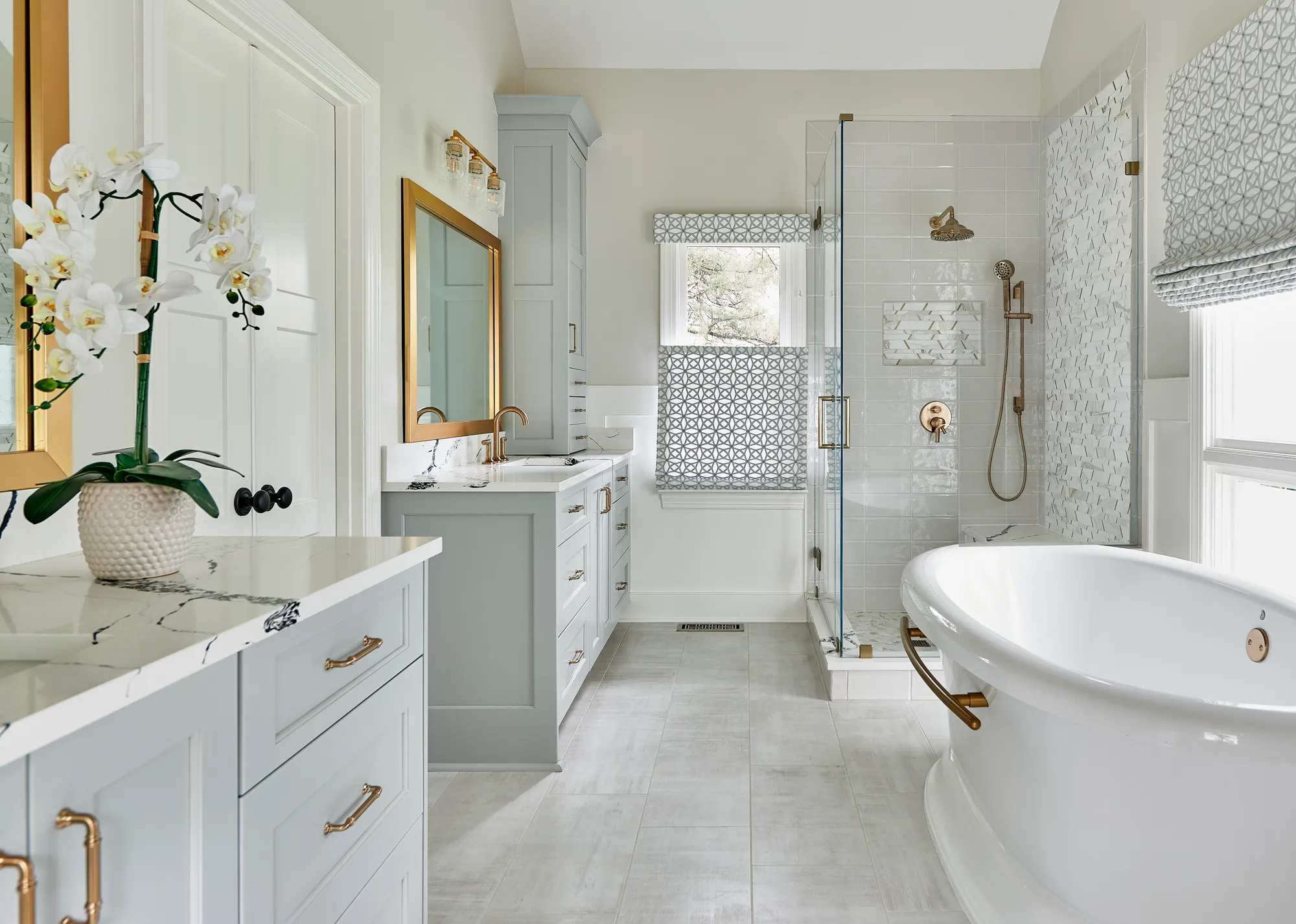 A luxurious bathroom with light blue double vanity and marble counters, a white freestanding tub, and a glass shower stall featuring gold hardware.