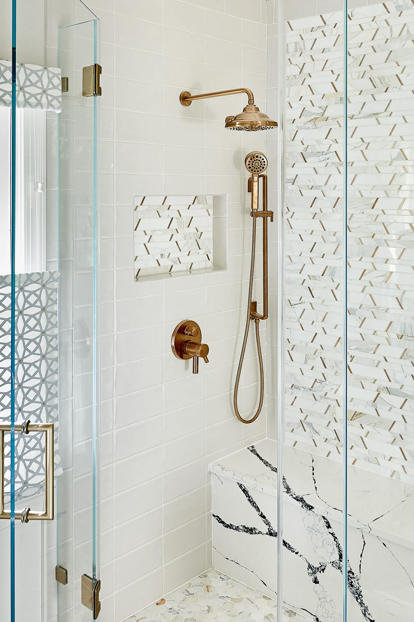 A contemporary shower stall with white tile, gold fixtures, a marble bench, and a striking feature wall of white and gold mosaic tile.
